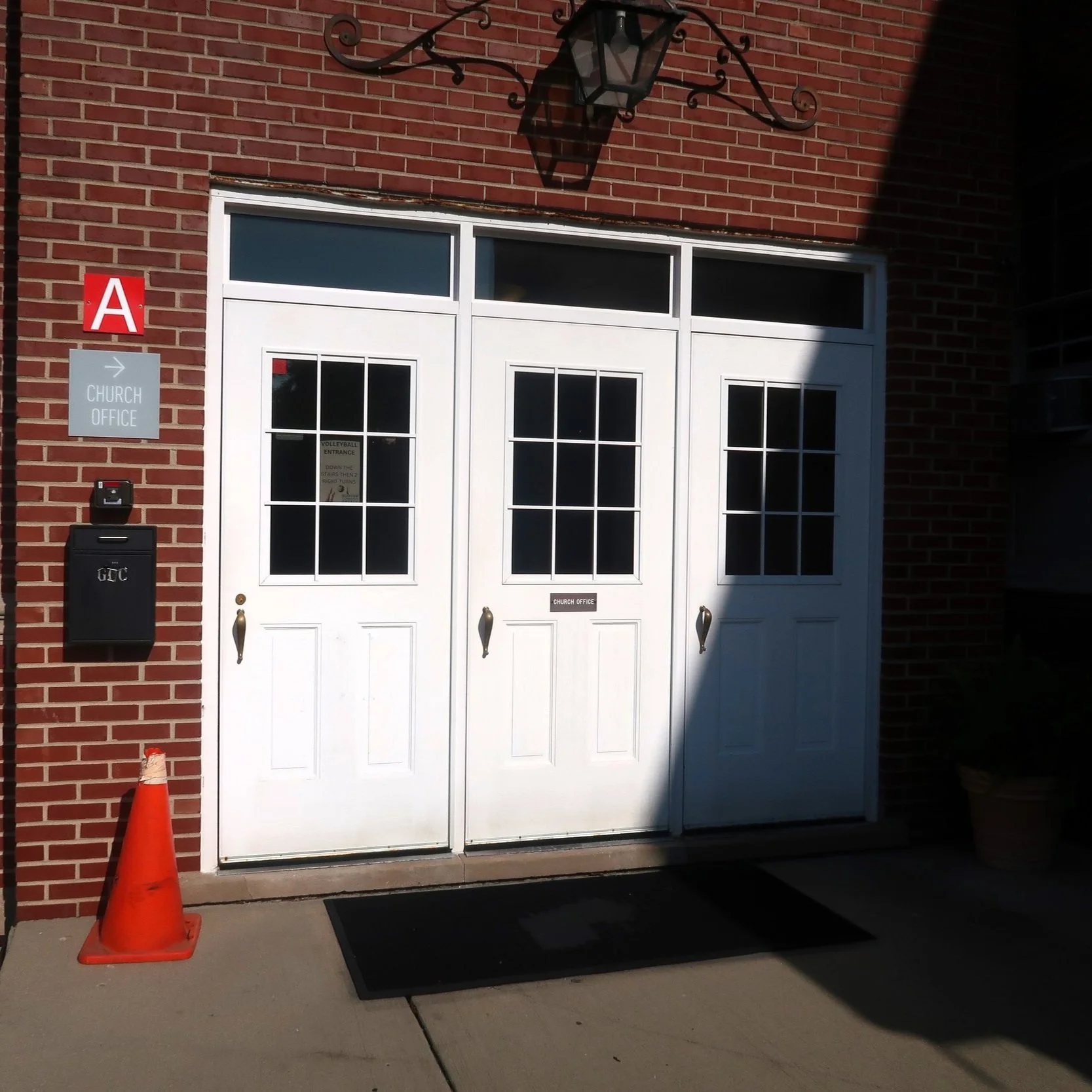 White double doors labeled 'Church Office' on a brick wall, with a black mailbox, a red traffic cone, and a sign pointing to the church office