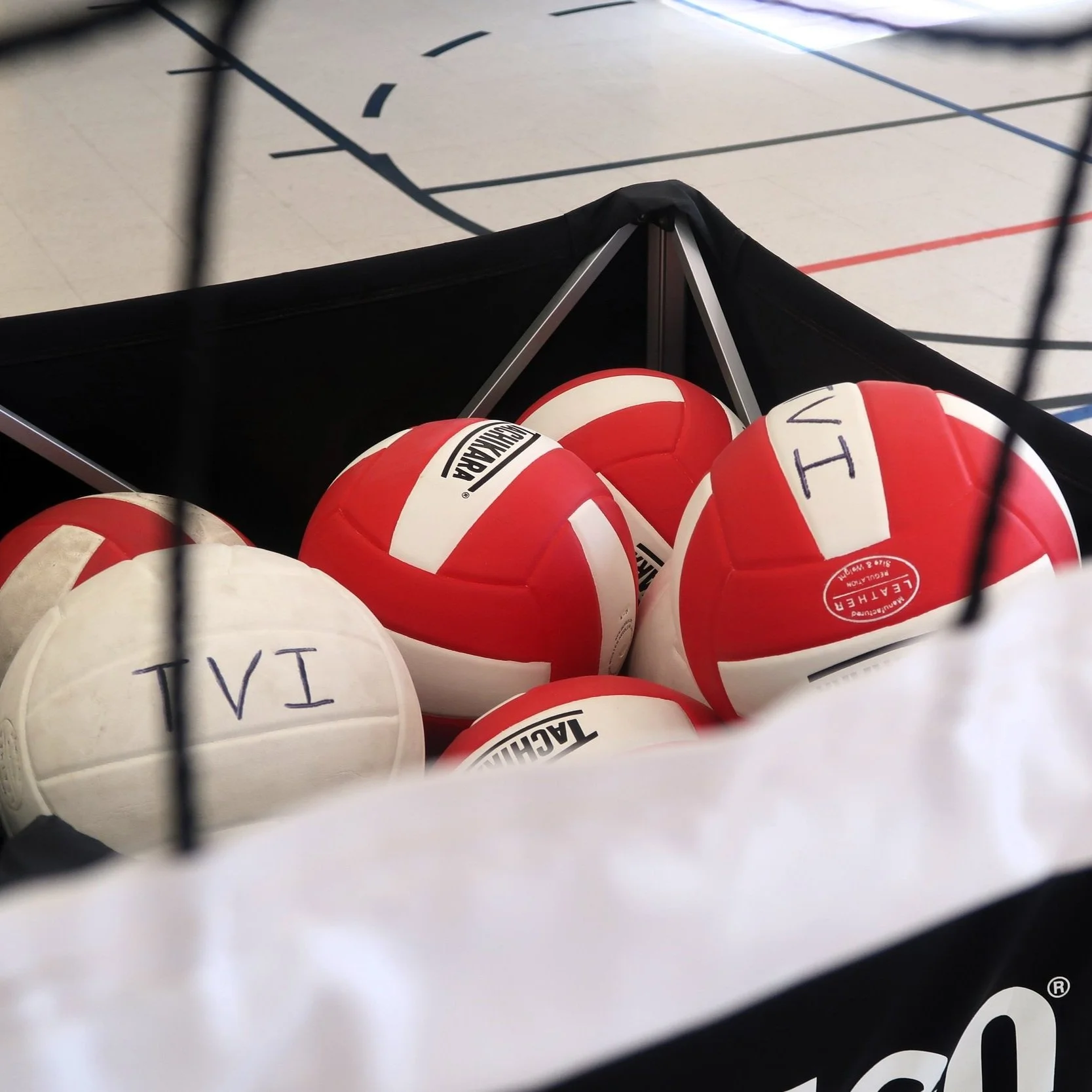 A black sports equipment bag containing several red and white volleyballs, some of which are marked with Roman numerals, inside a gymnasium with a basketball court in the background.
