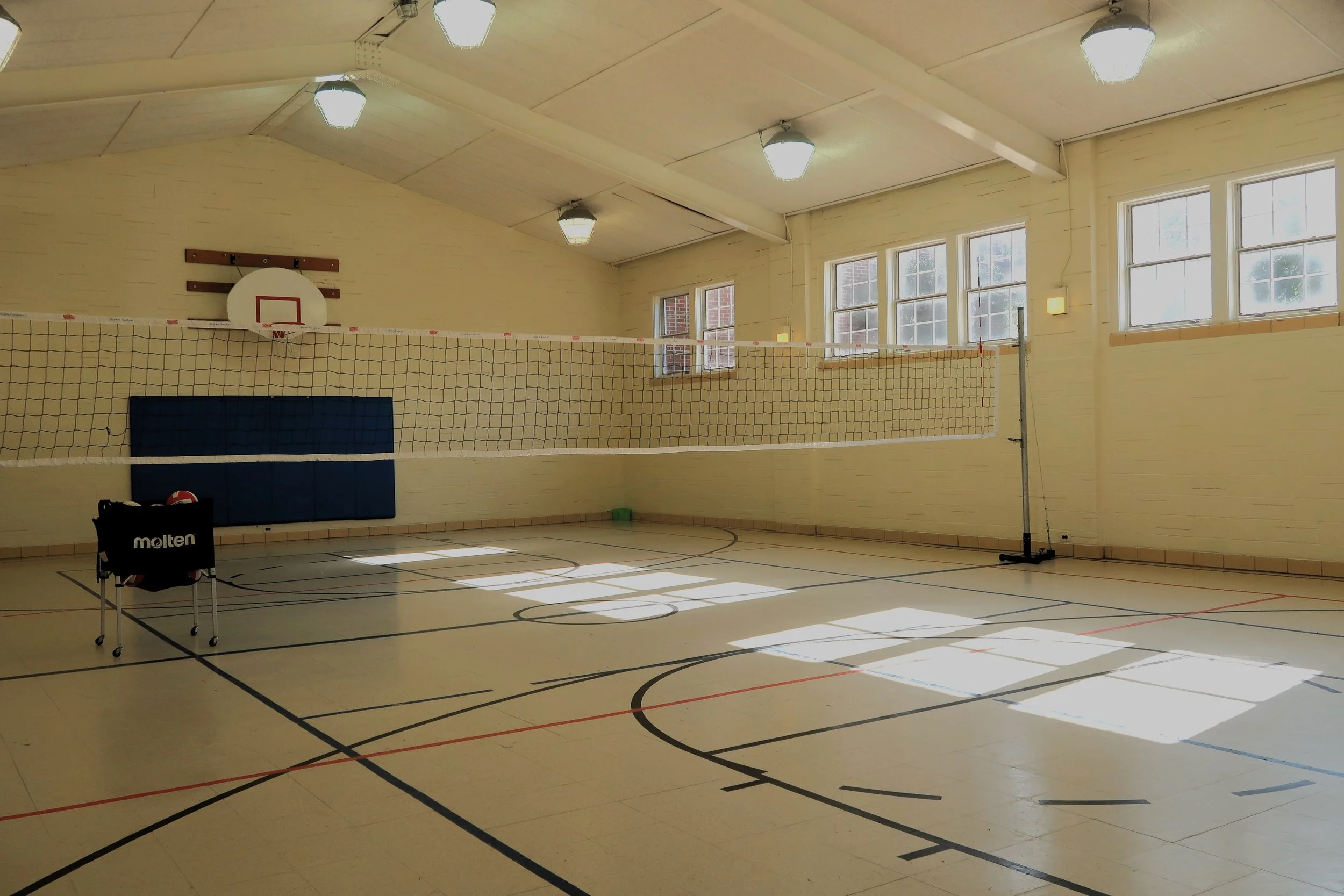 Empty indoor gymnasium with a volleyball net set up, basketball hoop, and sunlight streaming through multiple windows.