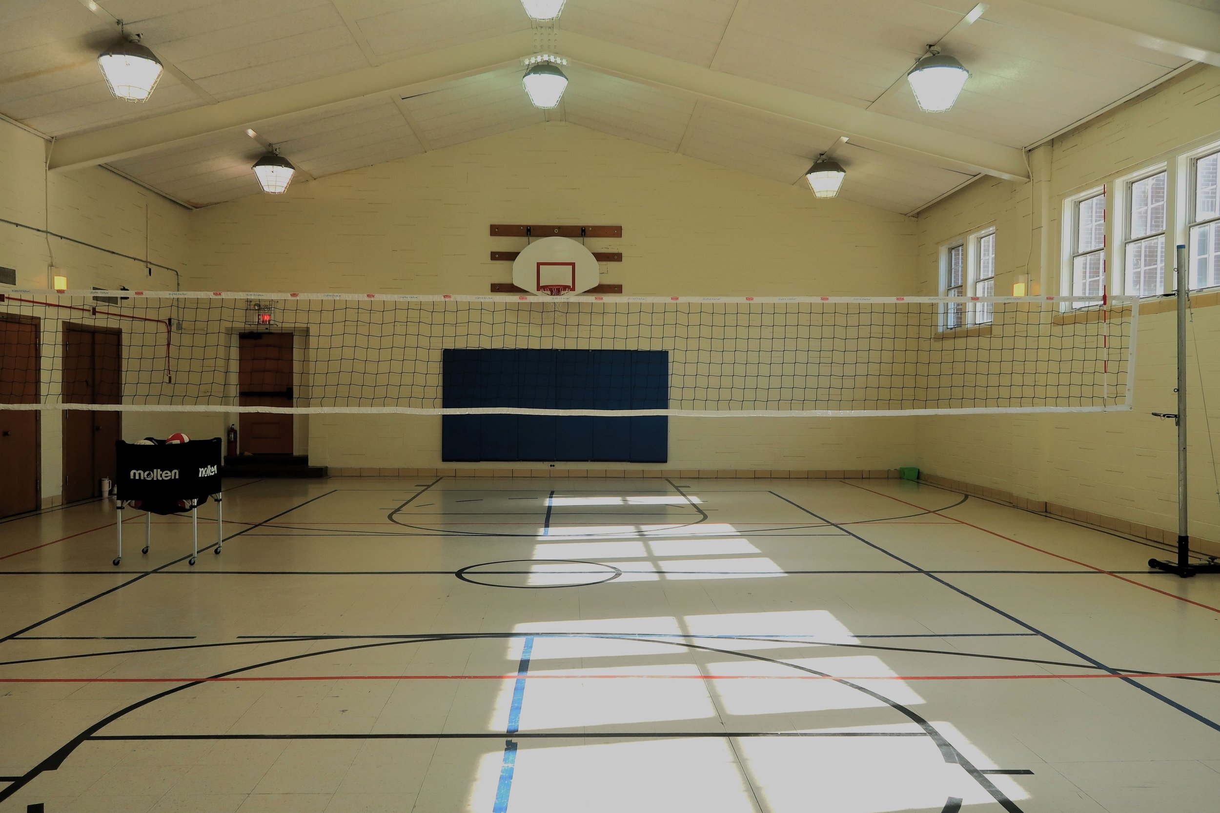 Empty indoor gymnasium with volleyball net, basketball hoop, and gym mats. Bright sunlight coming through windows on the right side.