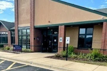 Exterior of a brick building with a green roof overhang, glass door entrance, and surrounding landscaping, located in a parking lot with a handicapped parking sign.
