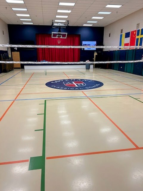 Empty gymnasium with volleyball net, U.S. Olympic logo on the floor, basketball hoop, and flags on the wall.