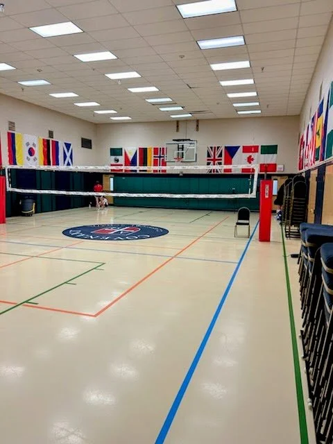 An indoor volleyball court with a volleyball net set up. Flags of various countries hang on the walls, and there are stacked chairs on the right side.