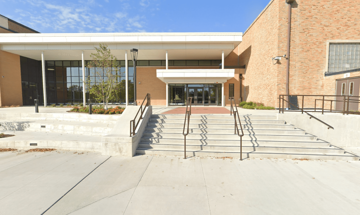 Front entrance of a modern school building with stairs, handrails, glass doors, and brick walls, under a blue sky.