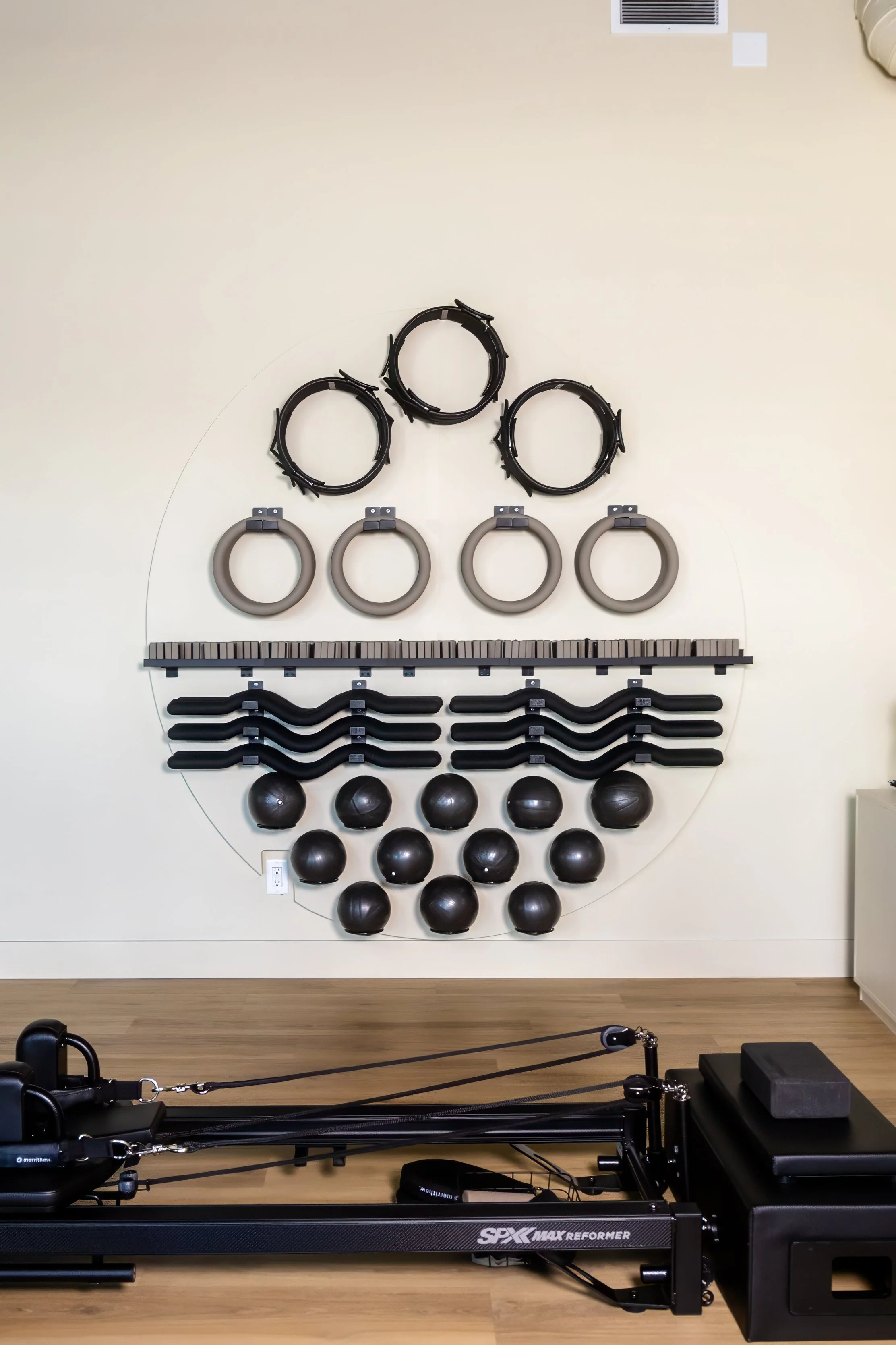 Wall with exercise equipment including black resistance balls, gray weighted circles, black wavy weighted bars, and black pilates rings, with a reformer machine in the foreground.