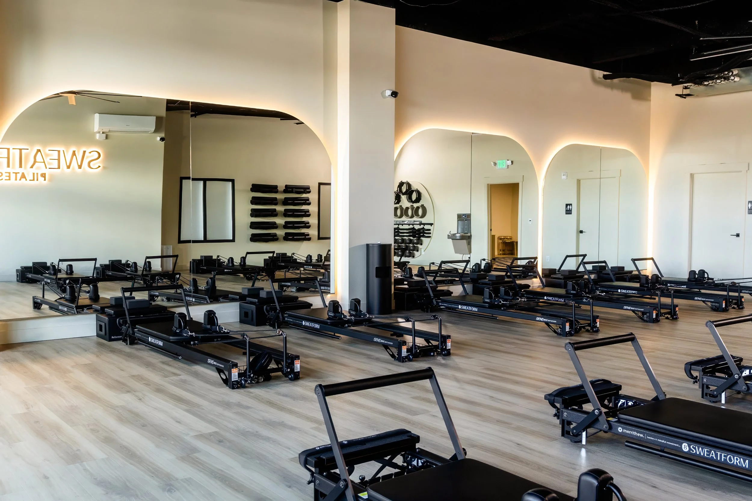 Pilates studio with reformer machines in a spacious, well-lit room with wooden floors, mirrors, and exercise equipment on the wall.