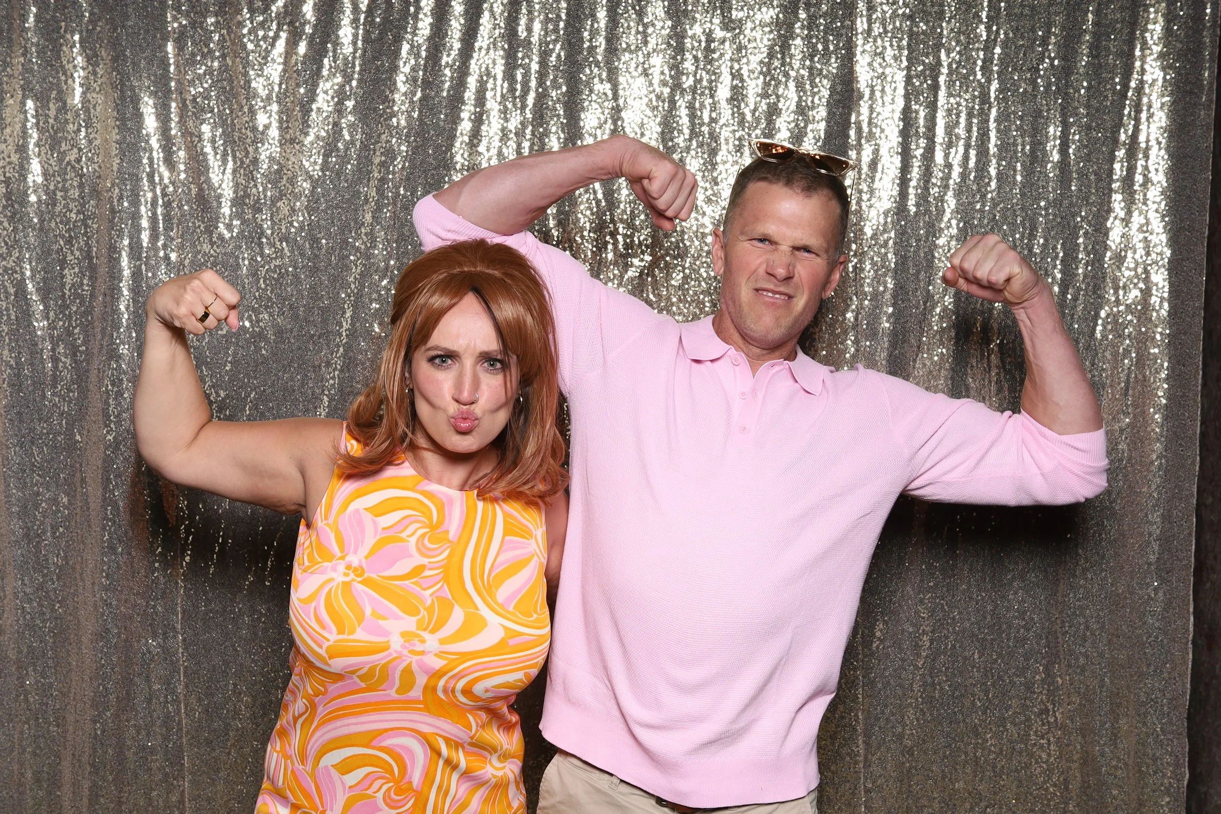 A woman with red hair in a colorful dress and a man in a pink shirt are posing in front of a silver glittery backdrop, both flexing their biceps and making a playful fierce face in a photo booth