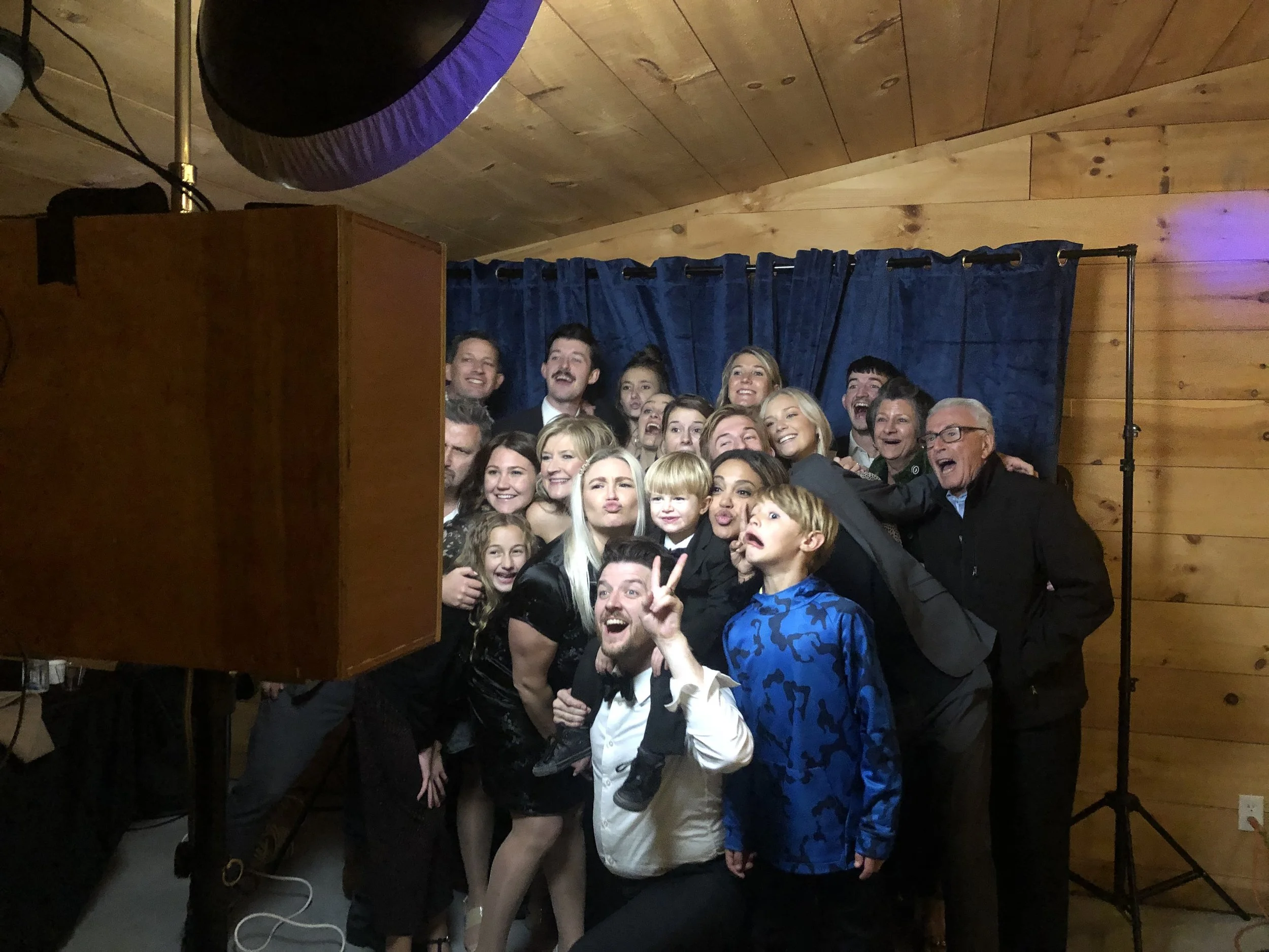 A large group of people, including children and adults, posing for a photo in front of a blue curtain backdrop in a wooden room, some making playful facial expressions and gestures, with a photographer and lighting equipment visible in a photo booth