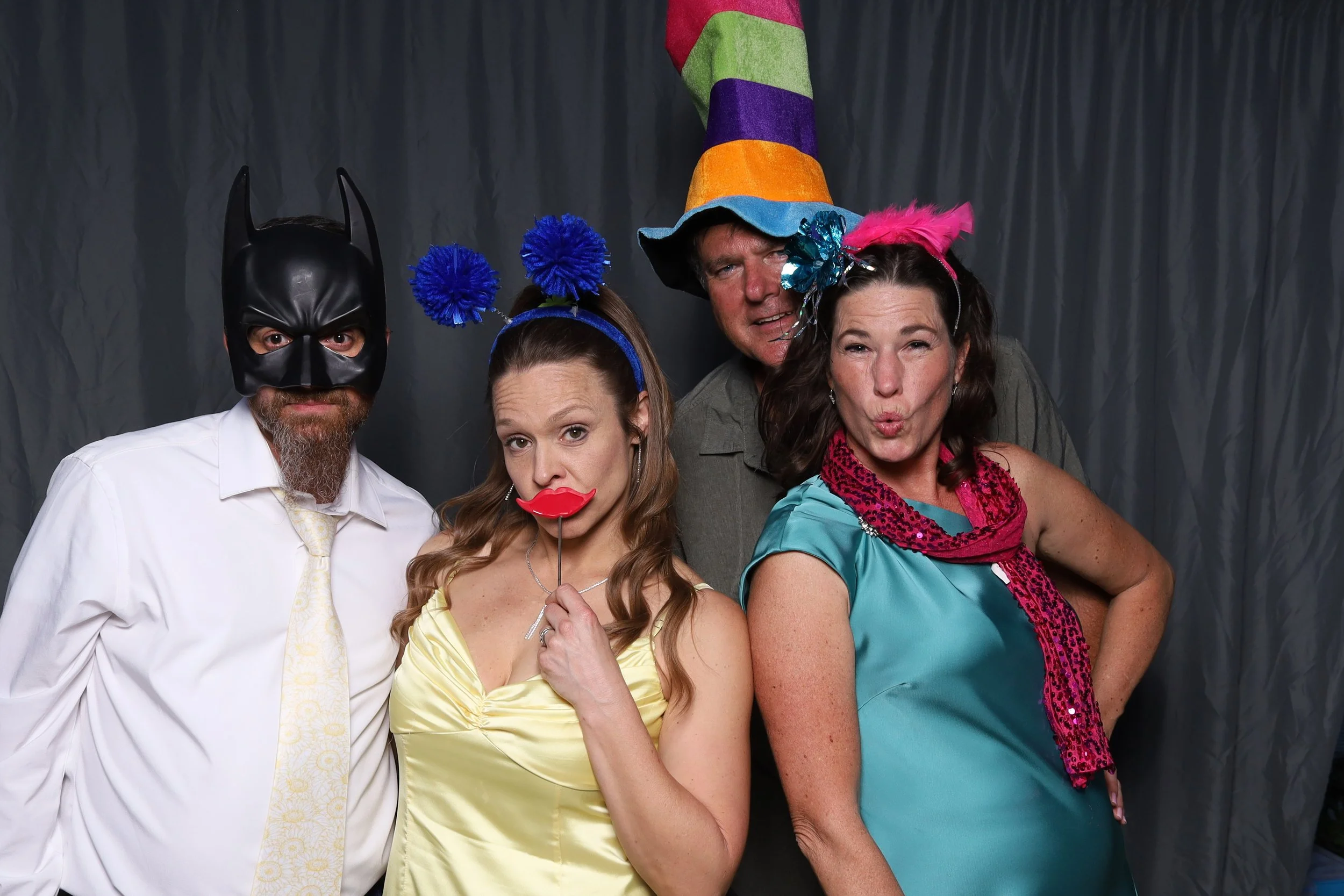 Group of four people dressed in colorful costumes and accessories, standing in front of a dark curtain backdrop in a photo booth.