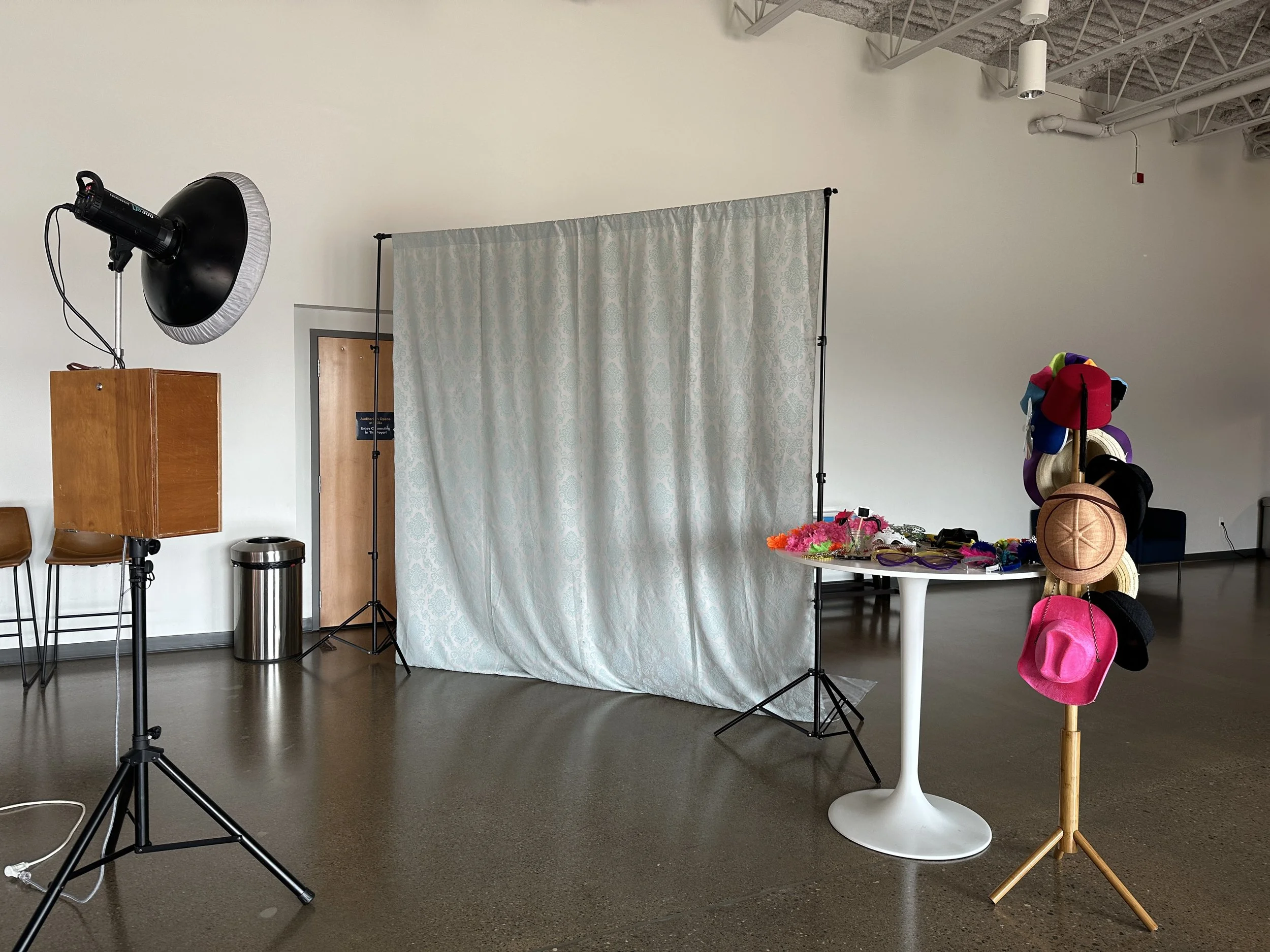 Photo of a photo booth setup with a white backdrop, a table with props including colorful feather boas and hats, and a hat stand with various hats.