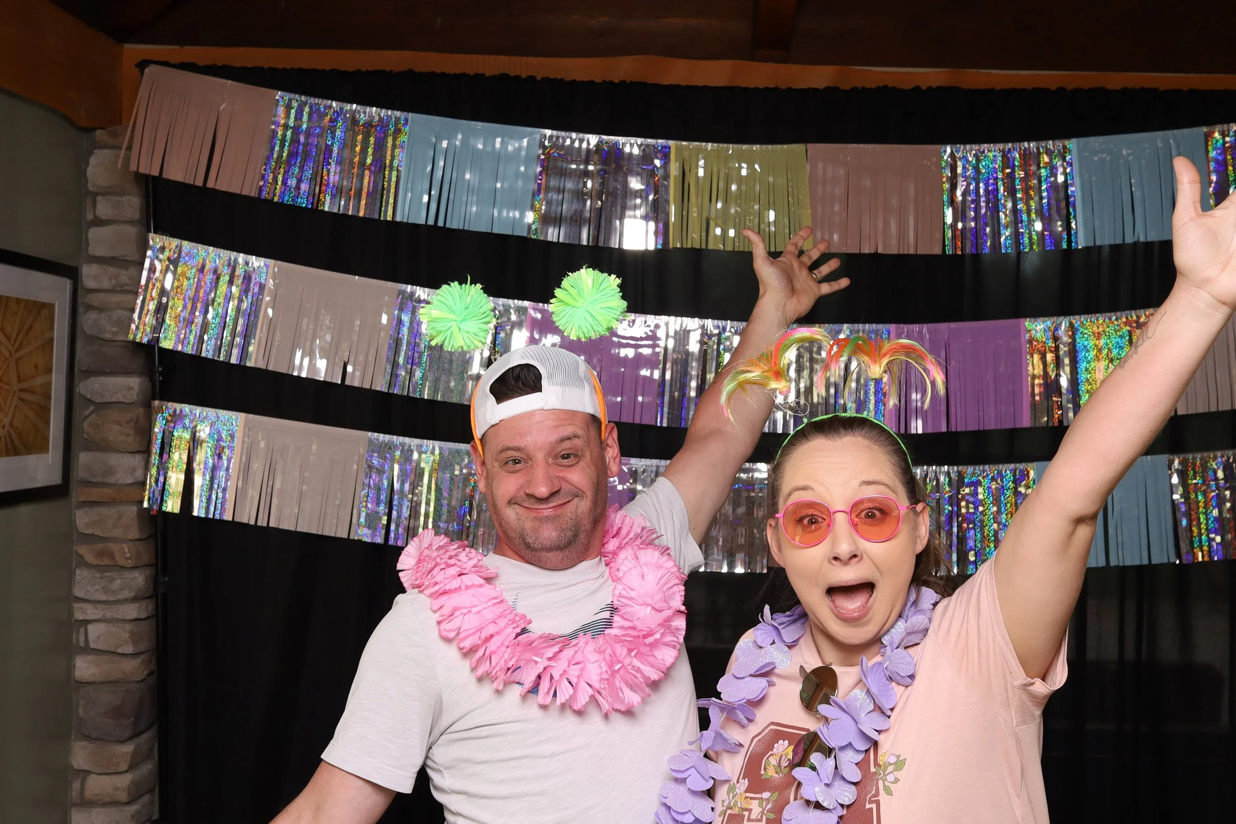 Two people celebrating at a party with festive decorations, wearing colorful leis, novelty glasses, and feathered hair accessories, with a shiny, multicolored backdrop.