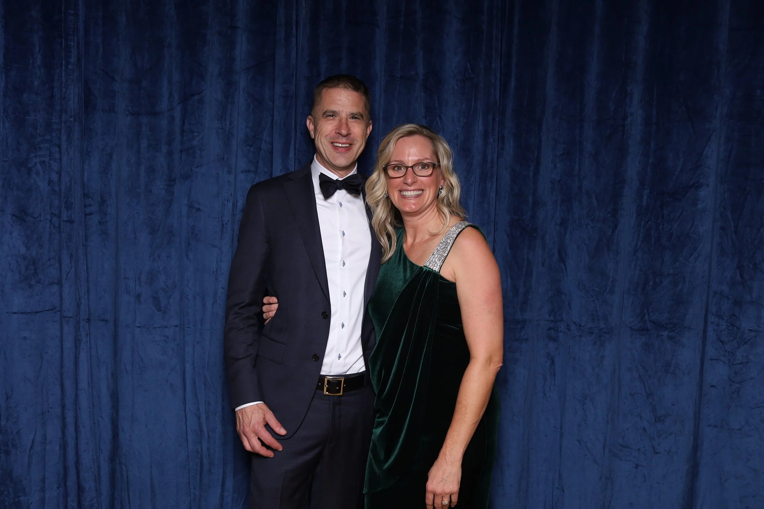 A man and a woman dressed in formal attire standing together in front of a blue curtain. The man is wearing a black tuxedo with a white shirt and black bow tie. The woman is wearing a green velvet dress with silver embellishments on the shoulder. The