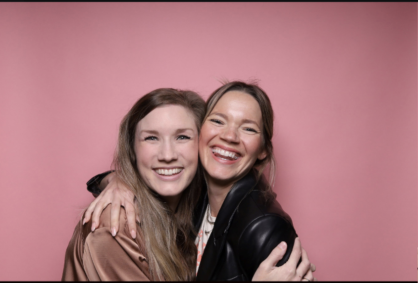 Two women smiling and hugging each other against a pink background.