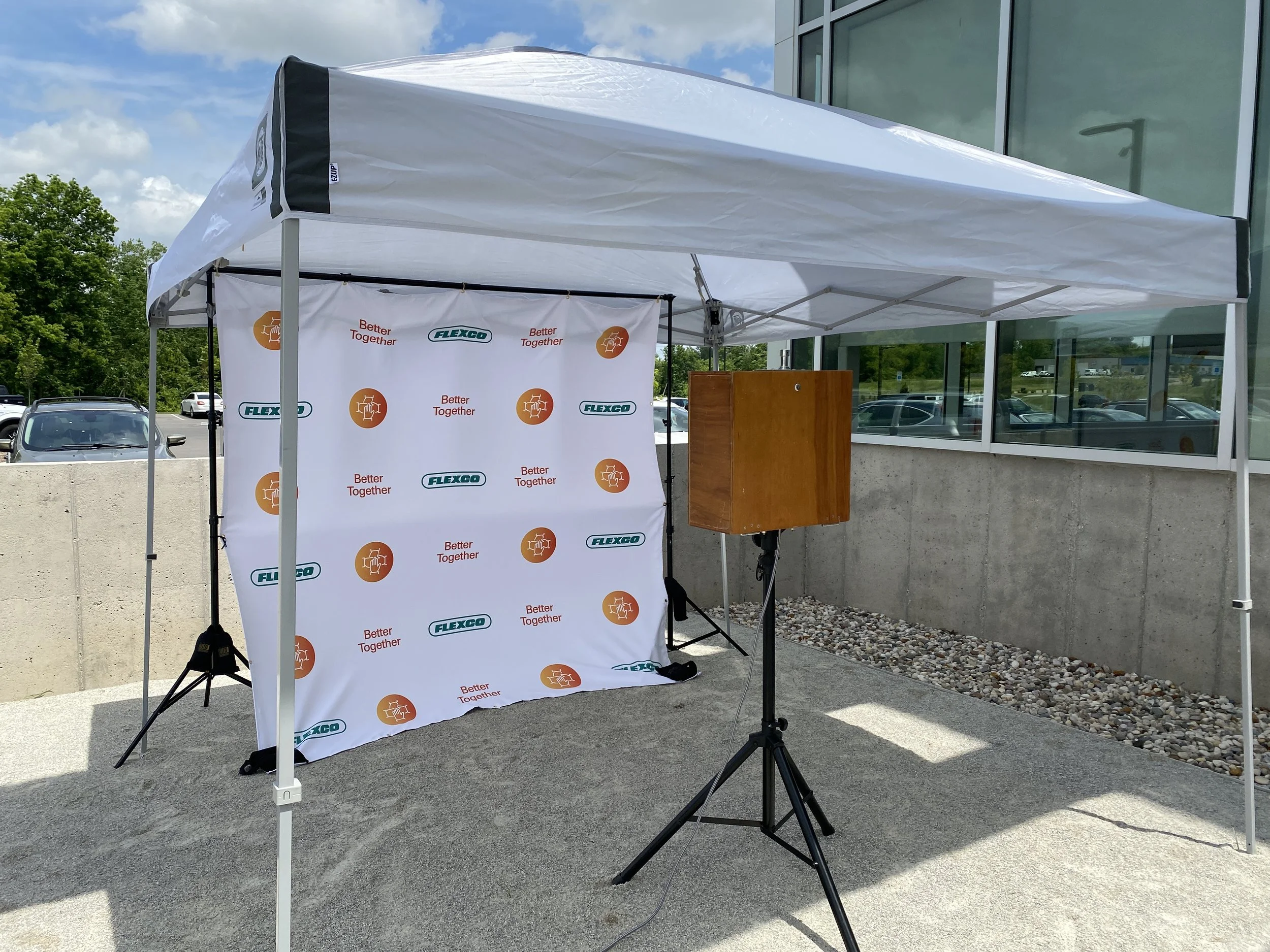 Outdoor event setup with a white canopy tent, a backdrop with logos and slogans, and a photo booth for a corporate event.