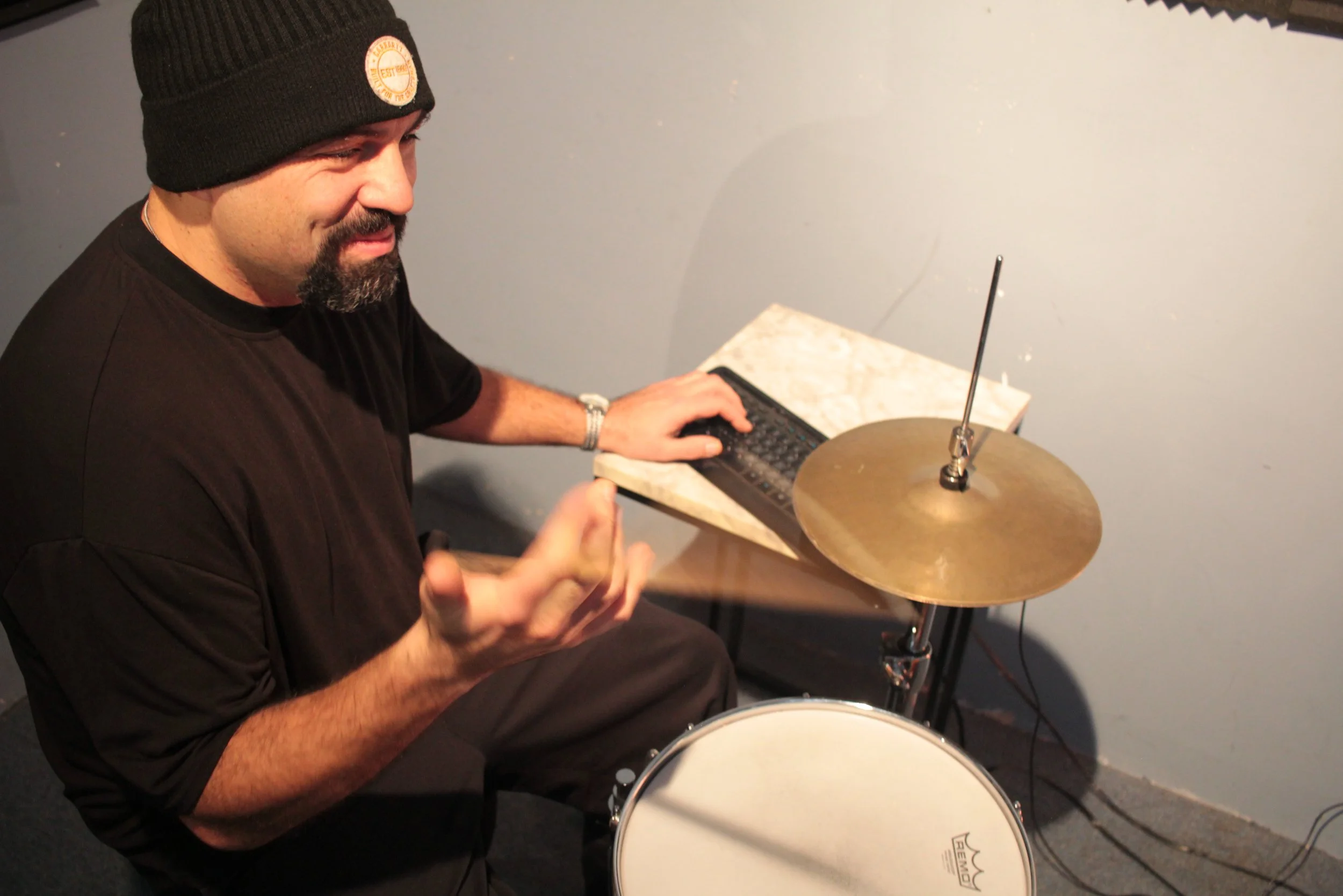 A man with a black beanie, black T-shirt, and a beard is sitting at a small drum set with a cymbal and a white drum. He is using a small electronic keyboard placed on a stand and is gesturing with his hand, possibly explaining or playing music.