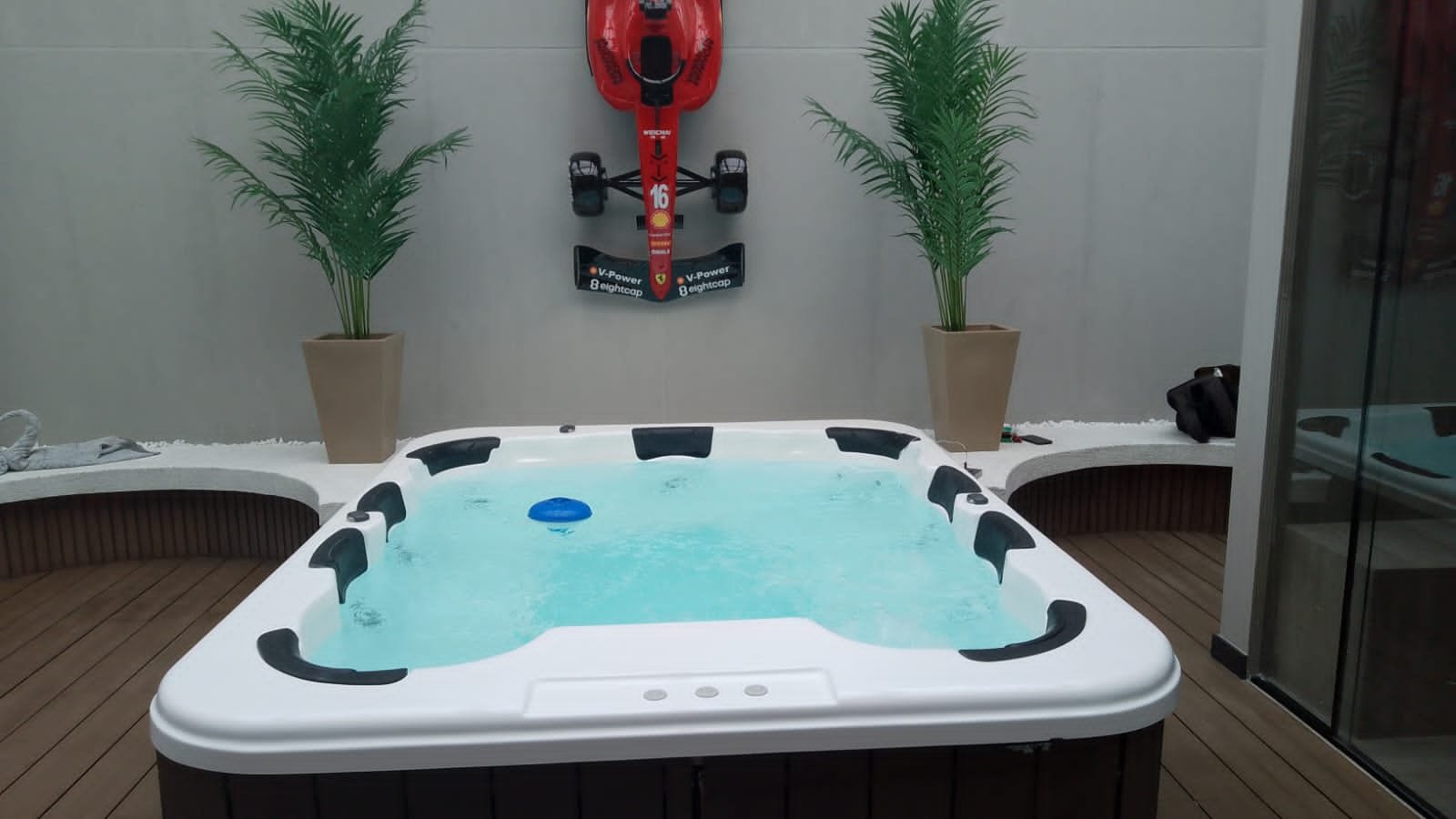 Jacuzzi con agua y burbujas en un área cerrada, decorada con dos plantas en macetas y una pared con un carrito de entrenamiento de carreras en la parte superior.