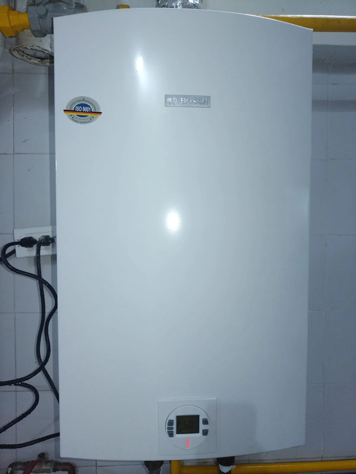 Calentador de agua blanco de la marca Bosch instalado en una pared, con pantalla digital y controles, conectado con tuberías y cables visibles a la izquierda y parte superior.
