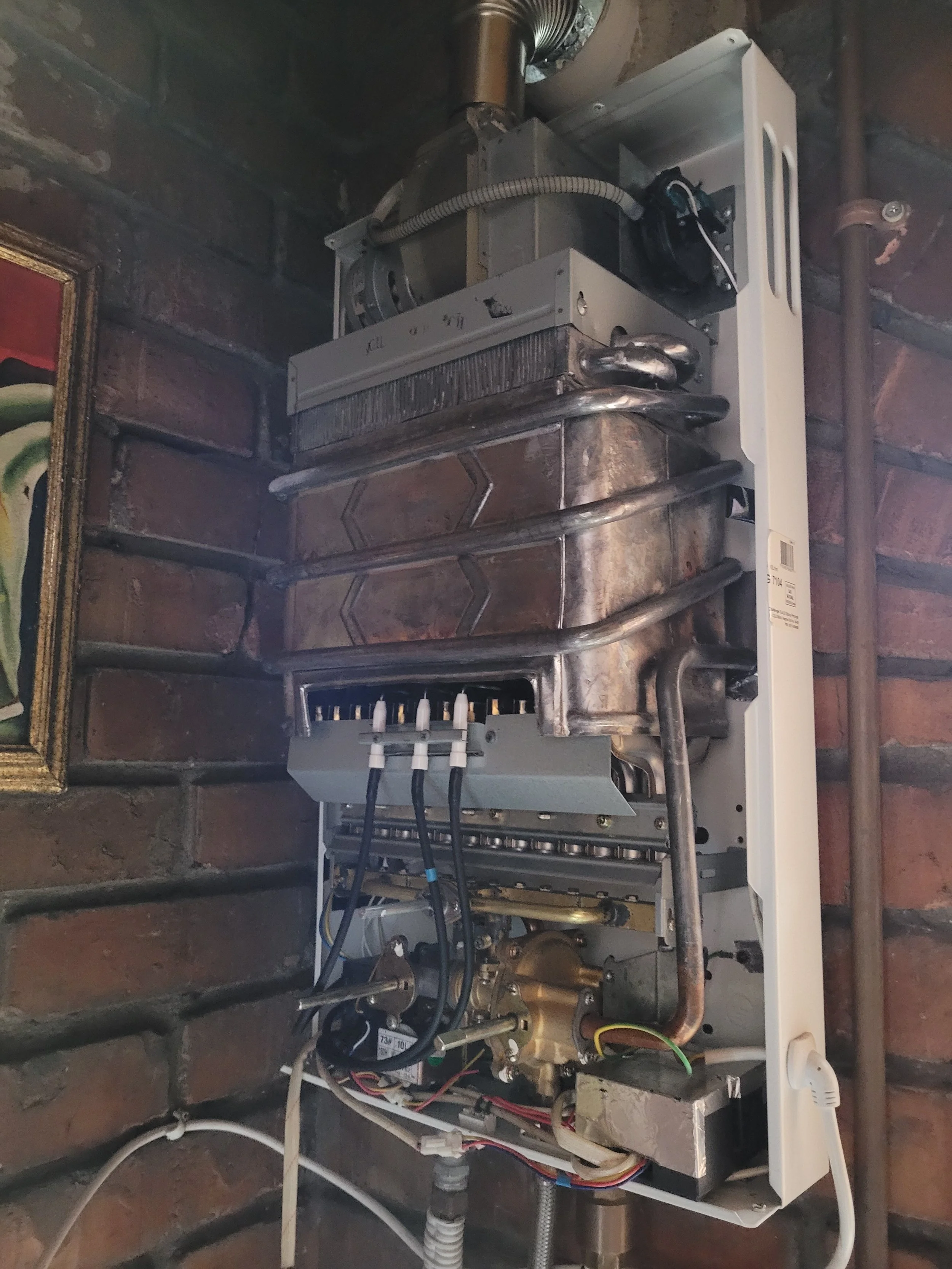 Vista de un calentador de agua con componentes internos visibles, montado en una pared de ladrillos con cables y tubos conectados.