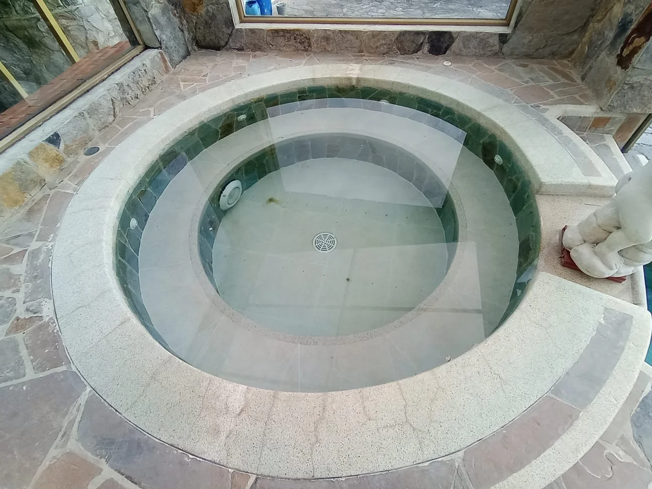 Jacuzzi de piedra con agua en un espacio interior con paredes de piedra y ventanas grandes.