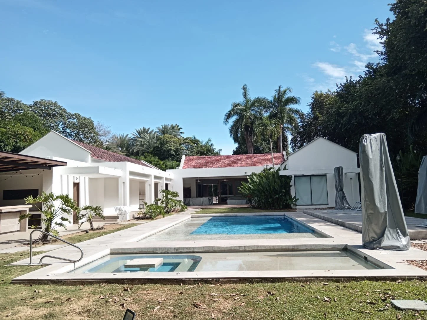 Amplia vista de una piscina dentro de una casa moderna con paredes blancas y áreas verdes, rodeada de palmeras y otros árboles, bajo cielo despejado.