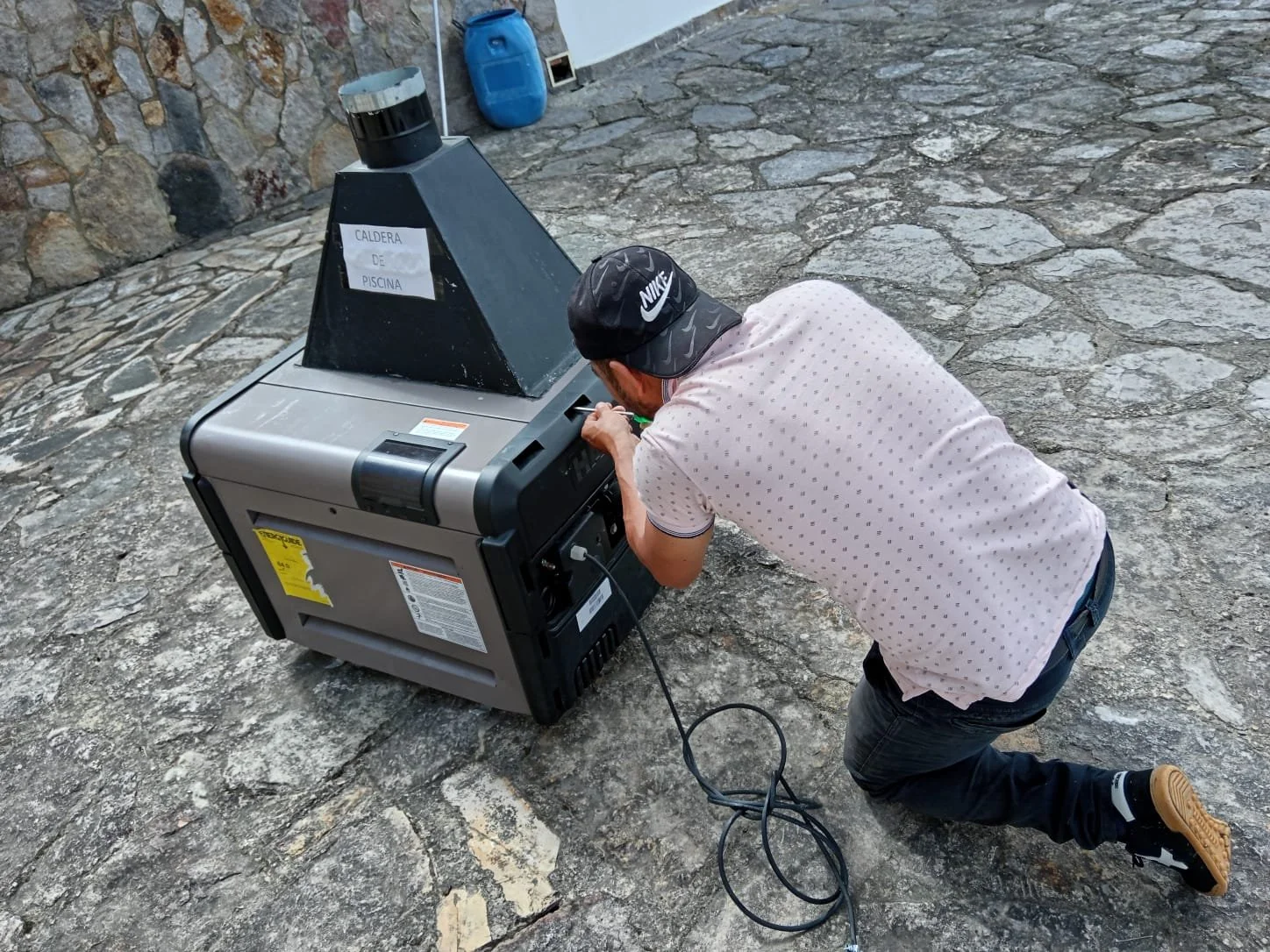 Un hombre arrodillado en el suelo ajustando una caldera de piscina en un espacio exterior pavimentado con piedras. En el fondo hay un muro de piedras y algunos recipientes azules.