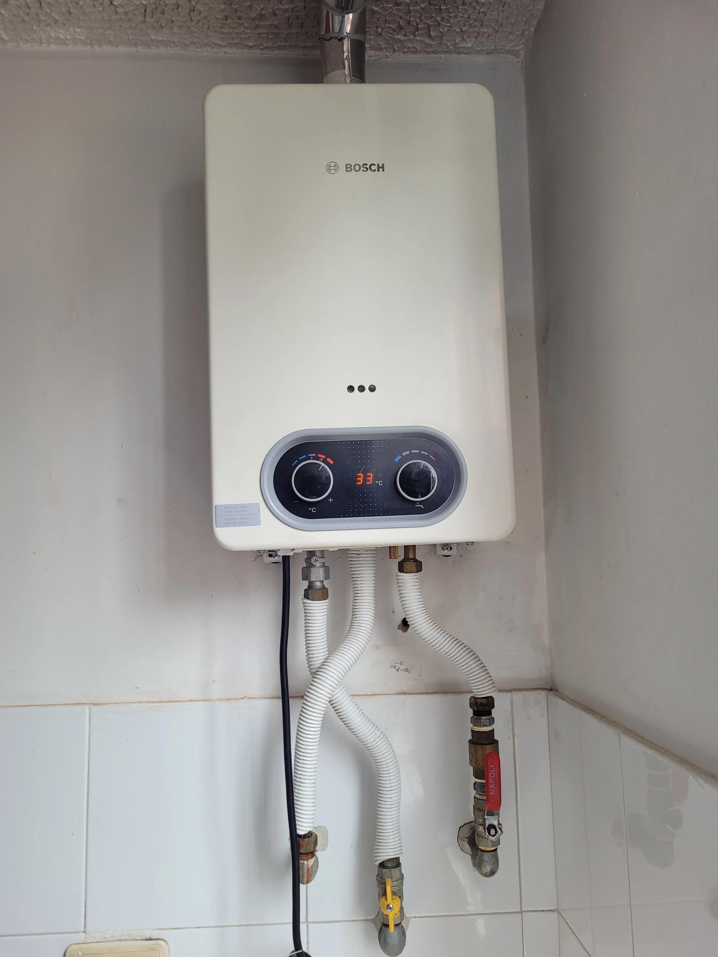 Calentador de agua Bosch montado en la pared con controles digitales y conexiones de plomería debajo, en una esquina de pared de azulejos.