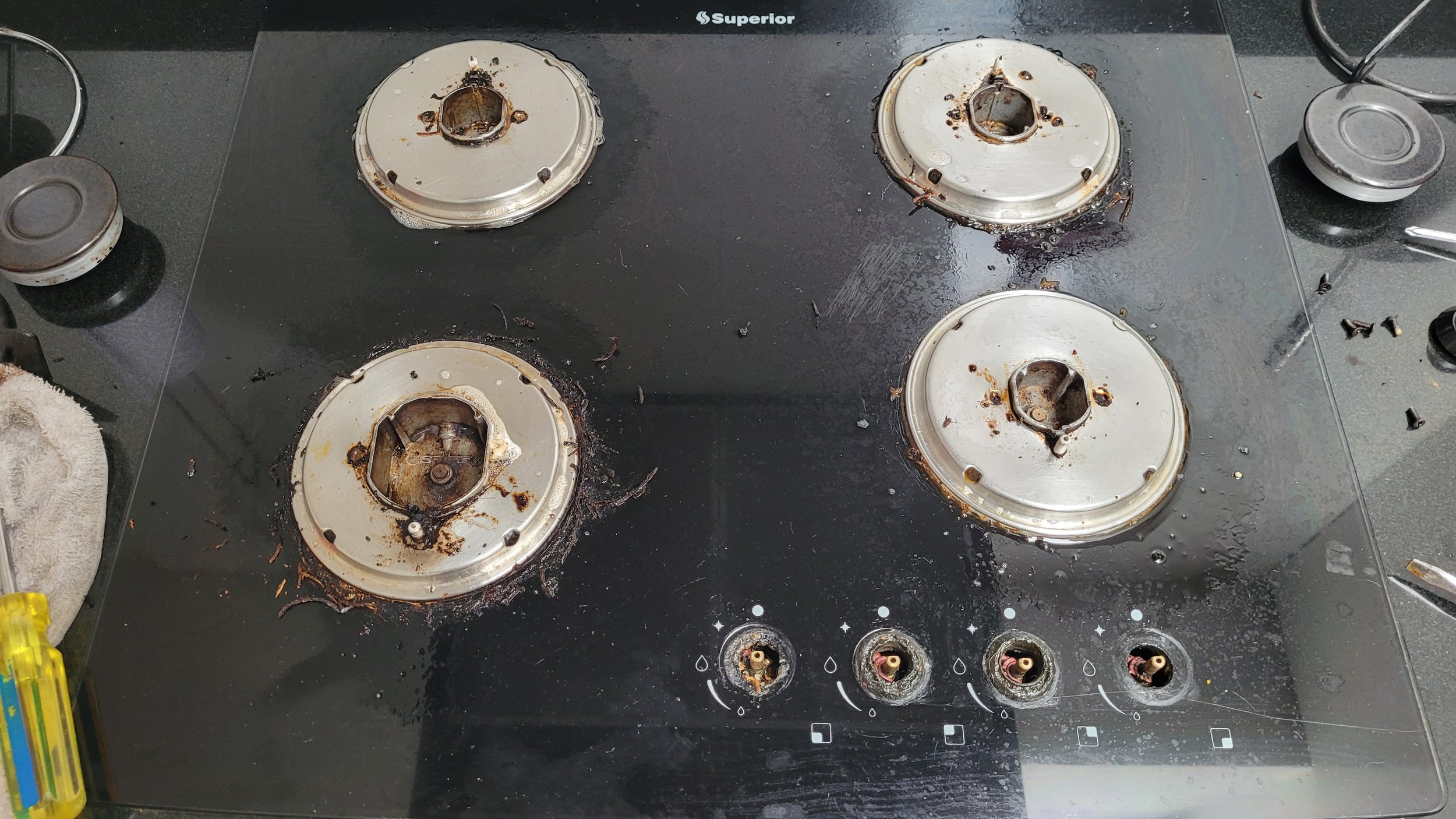 Estufa de cocina de vidrio con quemadores quemados y sucios, con tapas removidas y en proceso de limpieza.