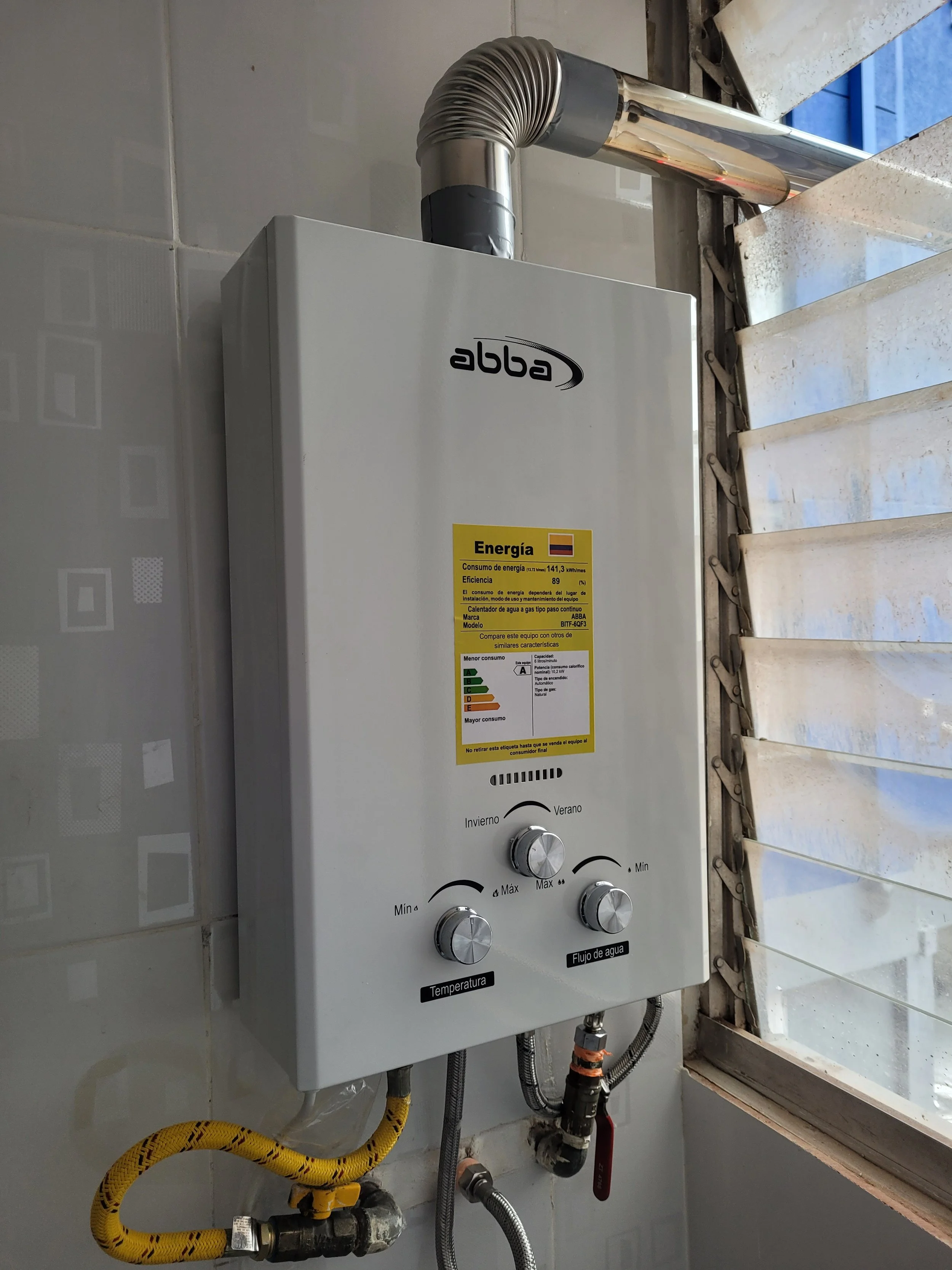 Calentador de agua instalado en una pared, con controles para ajustar la temperatura y el flujo de agua, y un tubo de escape de aluminio en la parte superior a la derecha.