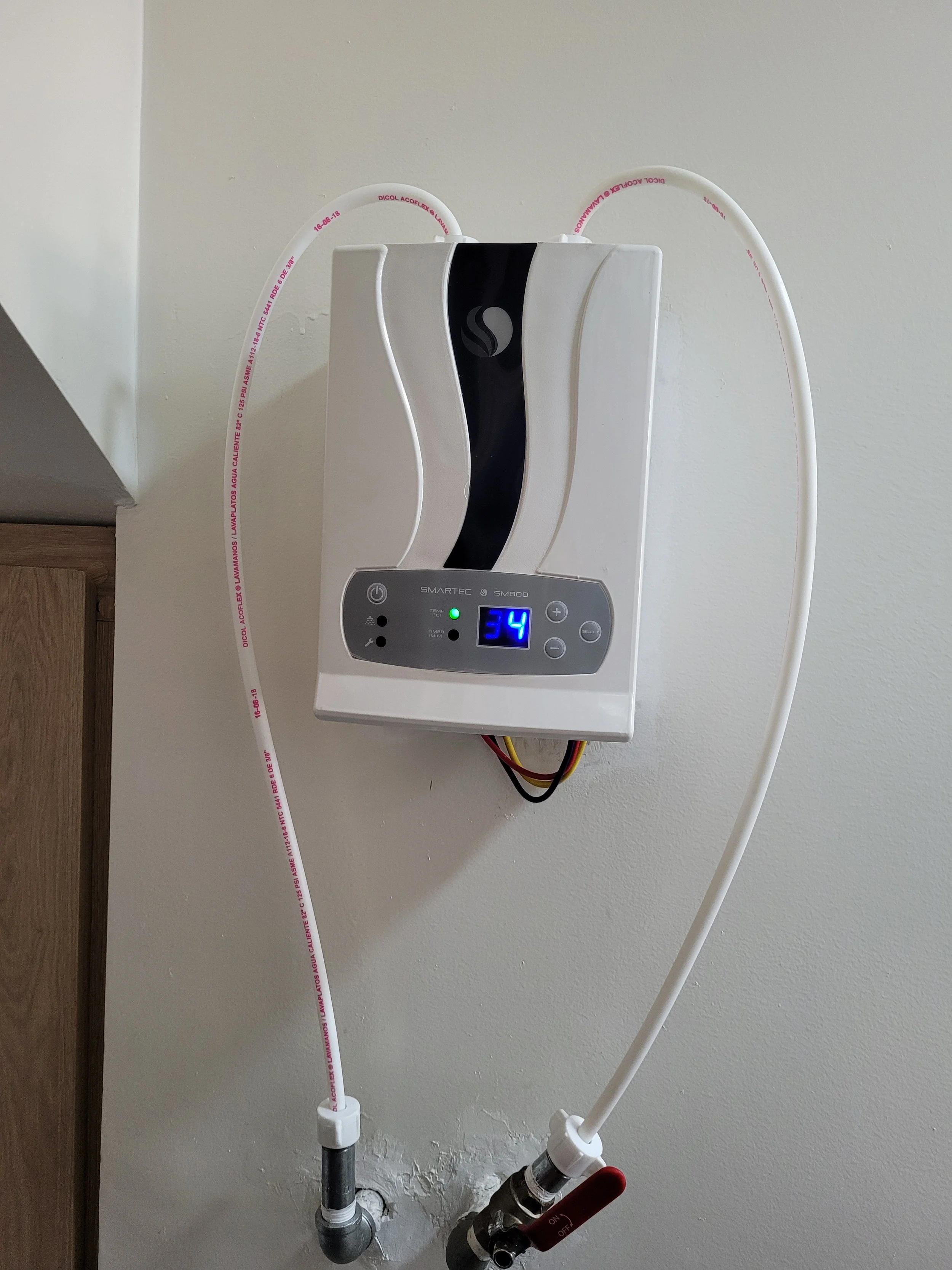 Calentador de agua eléctrico montado en la pared con tubos de entrada y salida, uno de los cuales está conectado a una tubería con válvula de cierre roja.