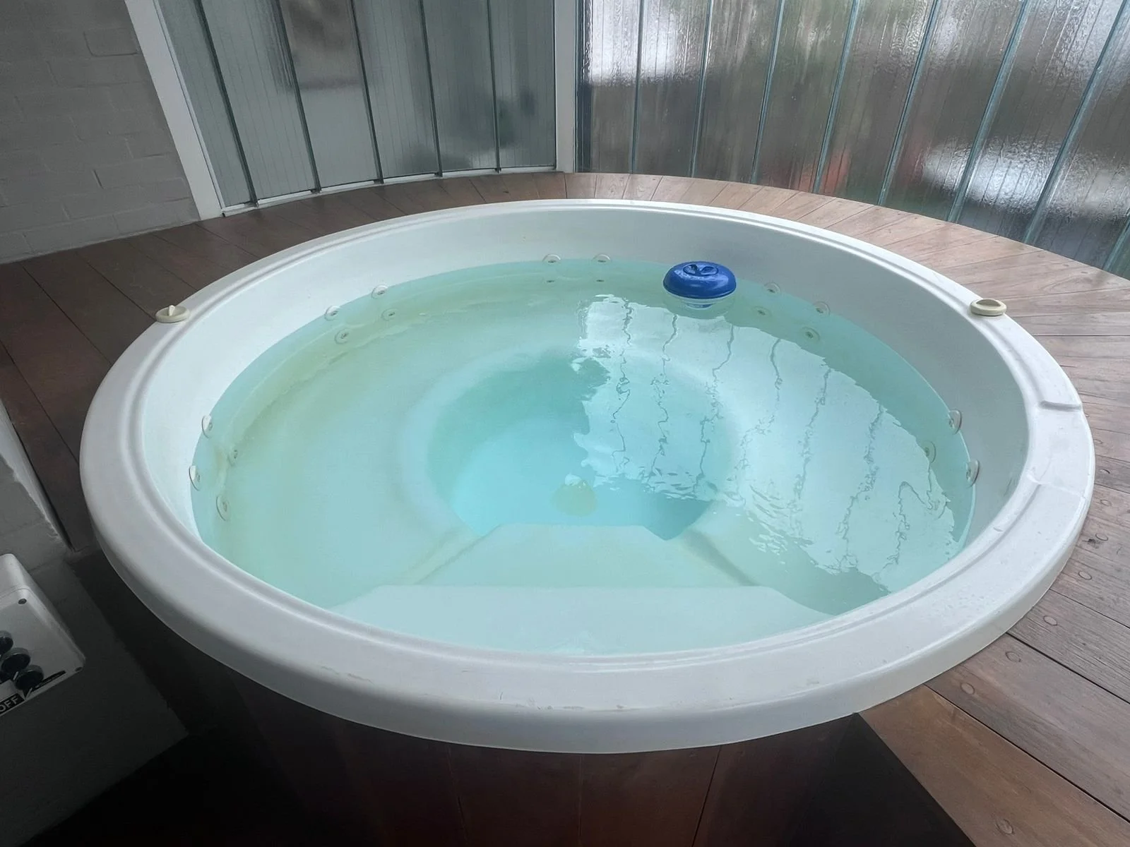 Una bañera de hidromasaje ovalada llena de agua, ubicada en una terraza de madera con paredes de vidrio y metal.