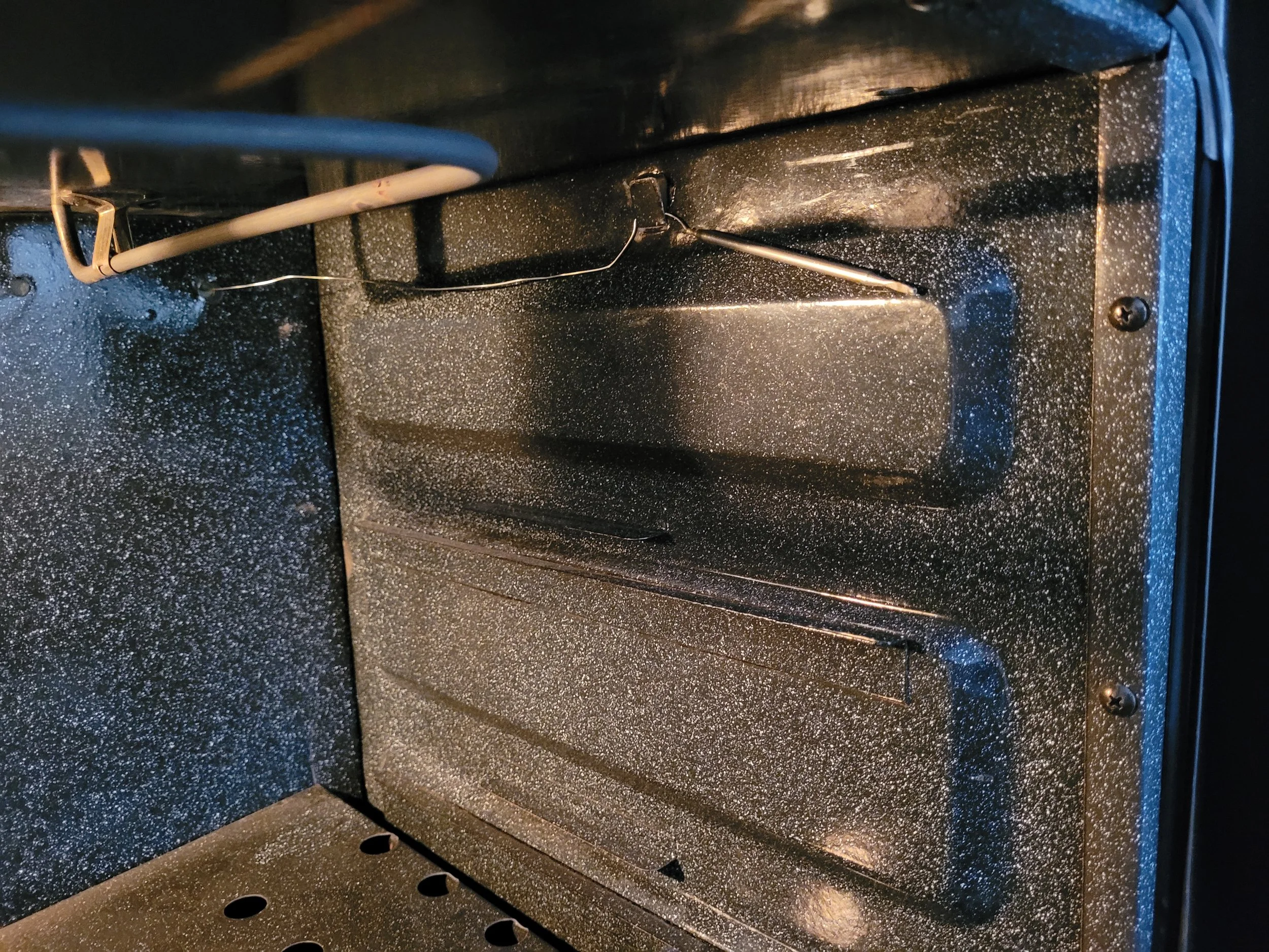 Interior de un horno con paredes metálicas grises con textura de puntos blancos, bandeja en la parte inferior y cables en la parte superior.