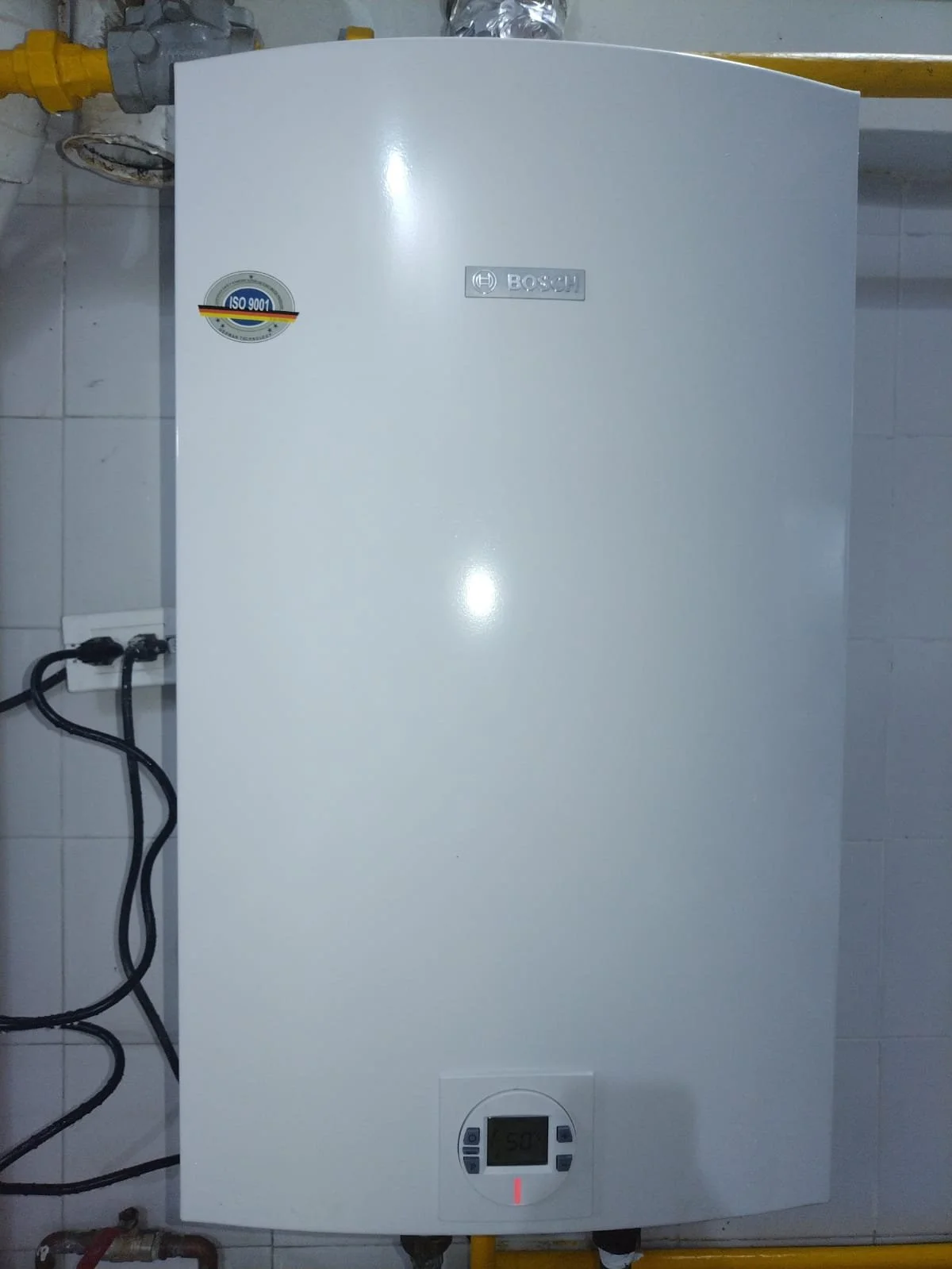 Calentador de agua Bosch en una pared con una pantalla digital y etiquetas, conectado por tuberías y cables en un espacio interior.
