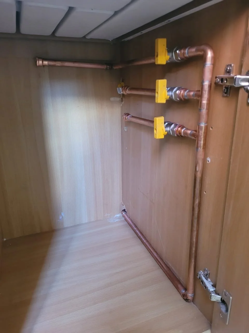 Sistema de tuberías de cobre con válvulas amarillas y conexiones metálicas en una pared de madera dentro de un mueble.