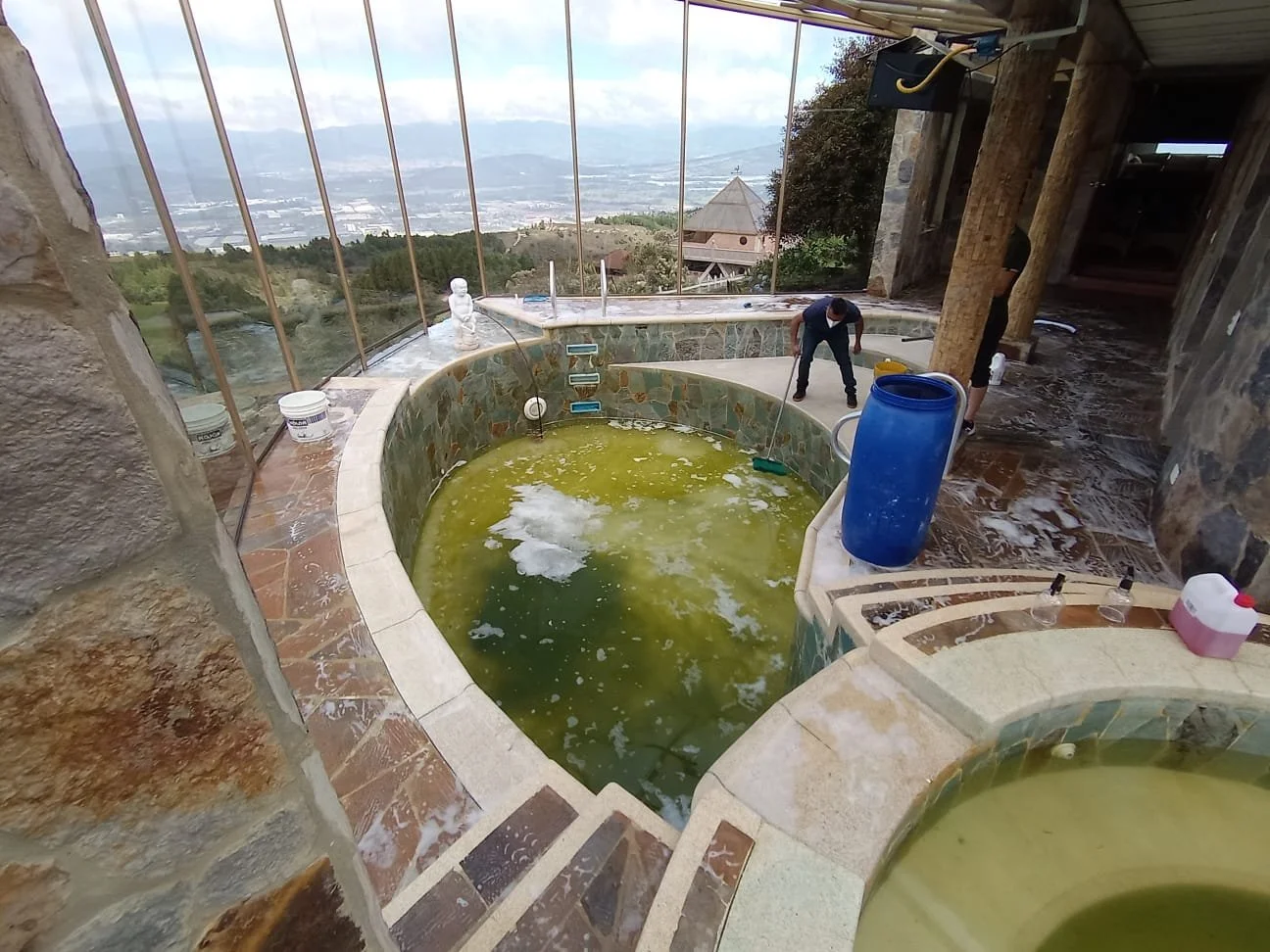 Piscina con agua contaminada y vegetación, en un entorno rústico y con vista panorámica de la ciudad y montañas, personas realizando tareas de limpieza.