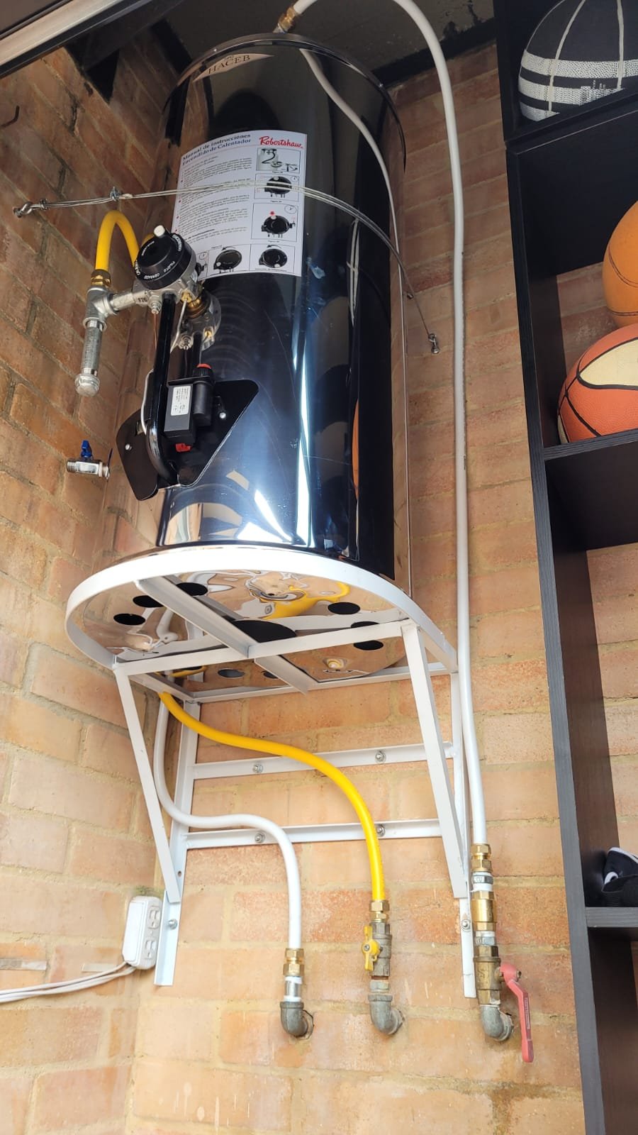 Un calentador de agua de cilindro negro instalado en la pared con conexiones de tubería y un manómetro en la parte superior, en un espacio con paredes de ladrillo.