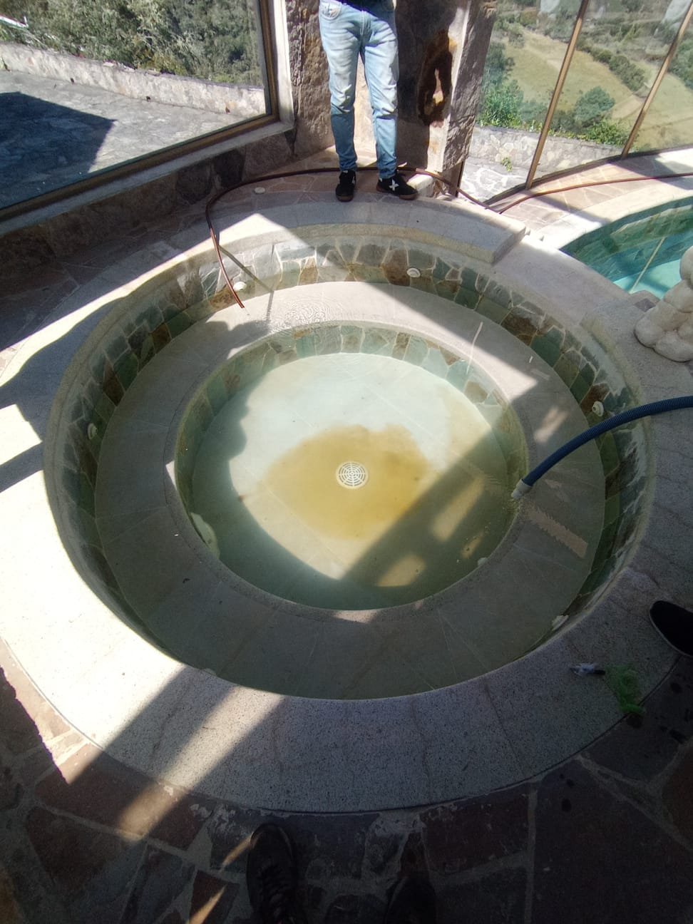 Piscina circular interior en construcción con agua y azulejos decorativos, una persona de jeans y zapatos negros en el fondo y gran ventanal con vista a vegetación.