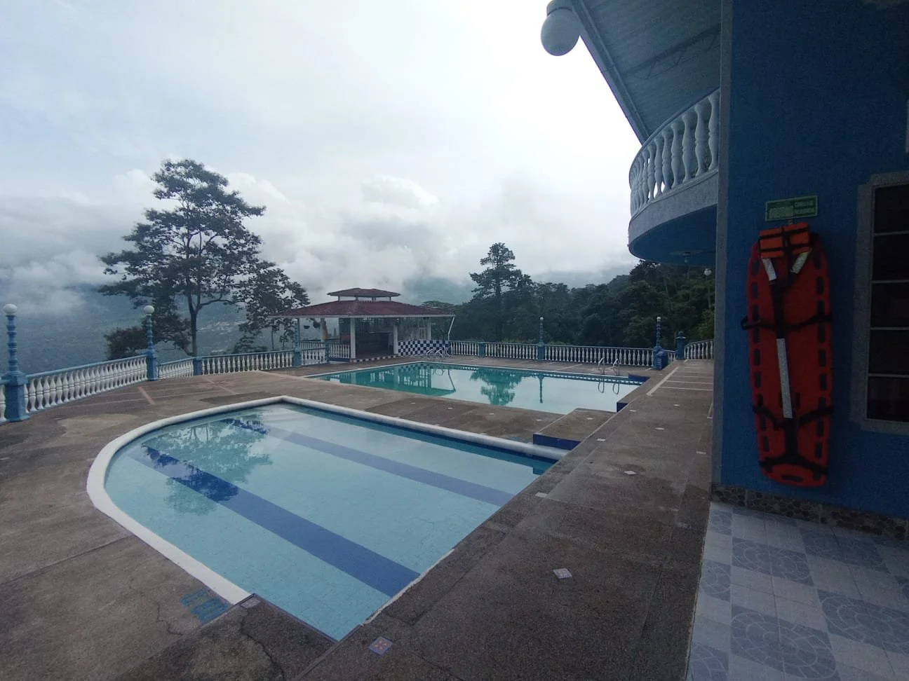 Piscina en un área elevada con vista a las montañas y árboles, edificio azul con balcón, colchoneta de rescatista colgada en la pared, cielo nublado.