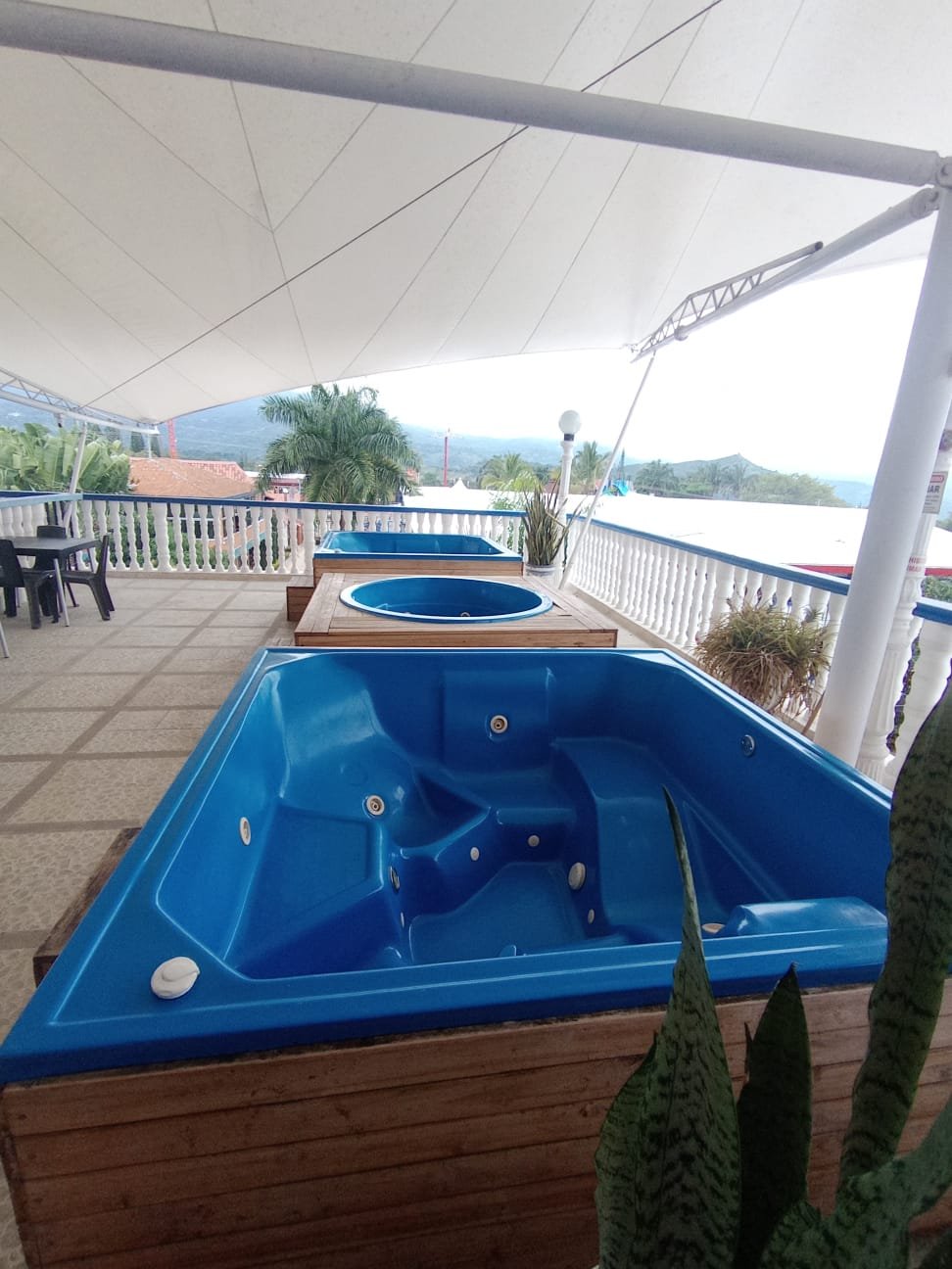 Jacuzzis en una terraza con techo de lona, vista a árboles y montañas, con mesas y sillas al fondo.