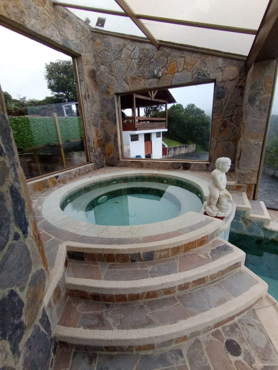 Una habitación con paredes de piedra, un jacuzzi en los escalones y una escultura de un niño sentado a un lado del jacuzzi. Ventanas grandes muestran un paisaje verde exterior.