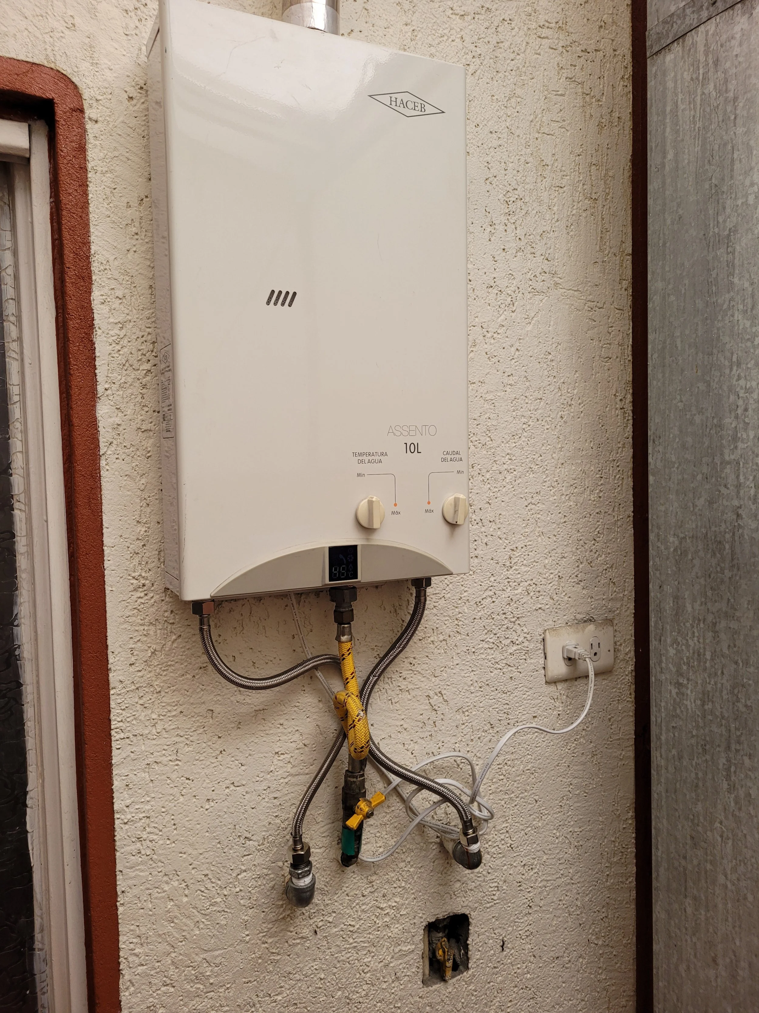 Calentador de agua montado en la pared con controles y conexiones eléctricas y de agua conectadas, en una pared de textura áspera.