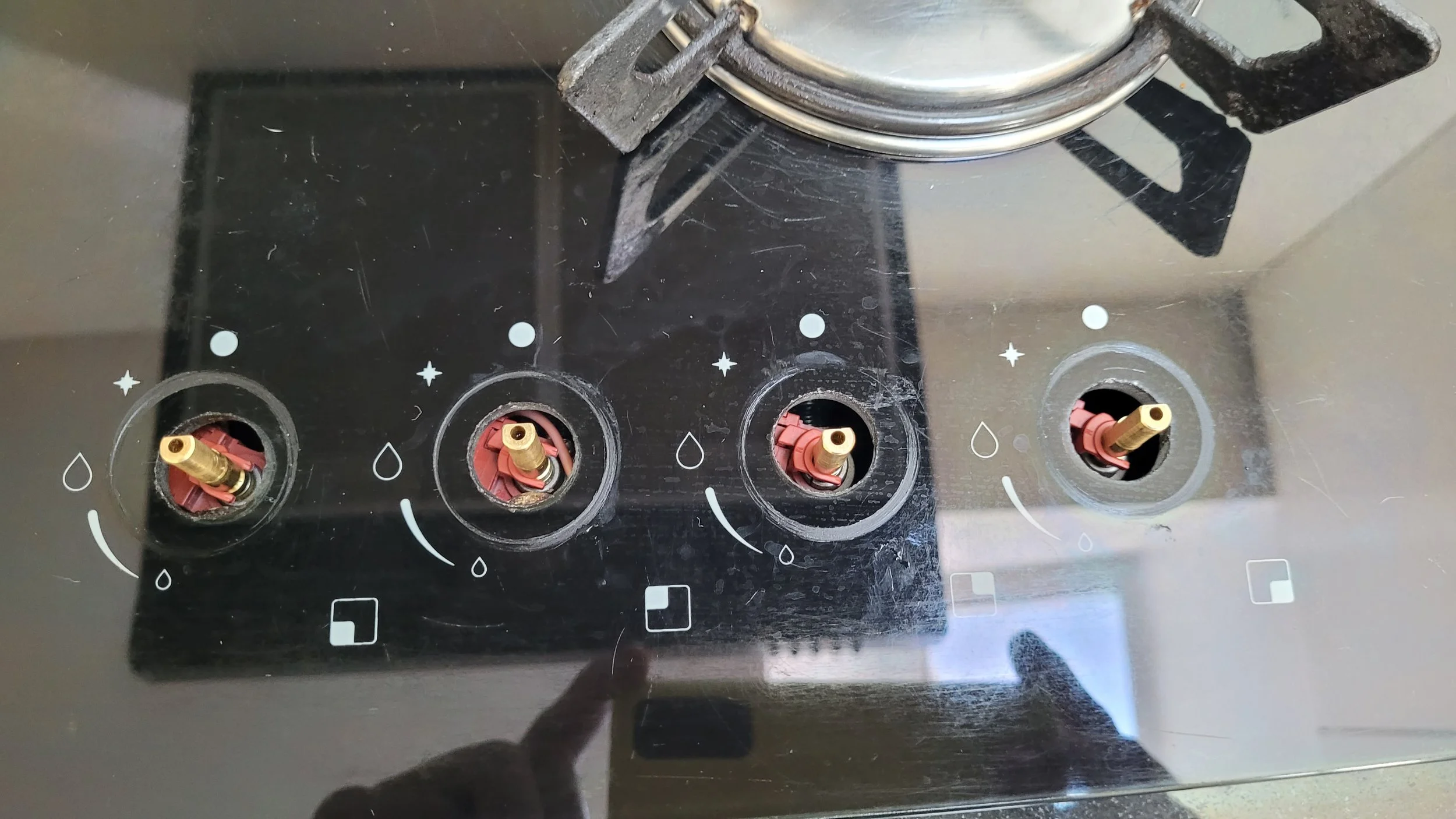 Superficie de cocción de estufa con cuatro quemadores, con tapa de cristal y marcas decorativas con gotas de agua y estrellas, y un litro de olla en uno de los quemadores.