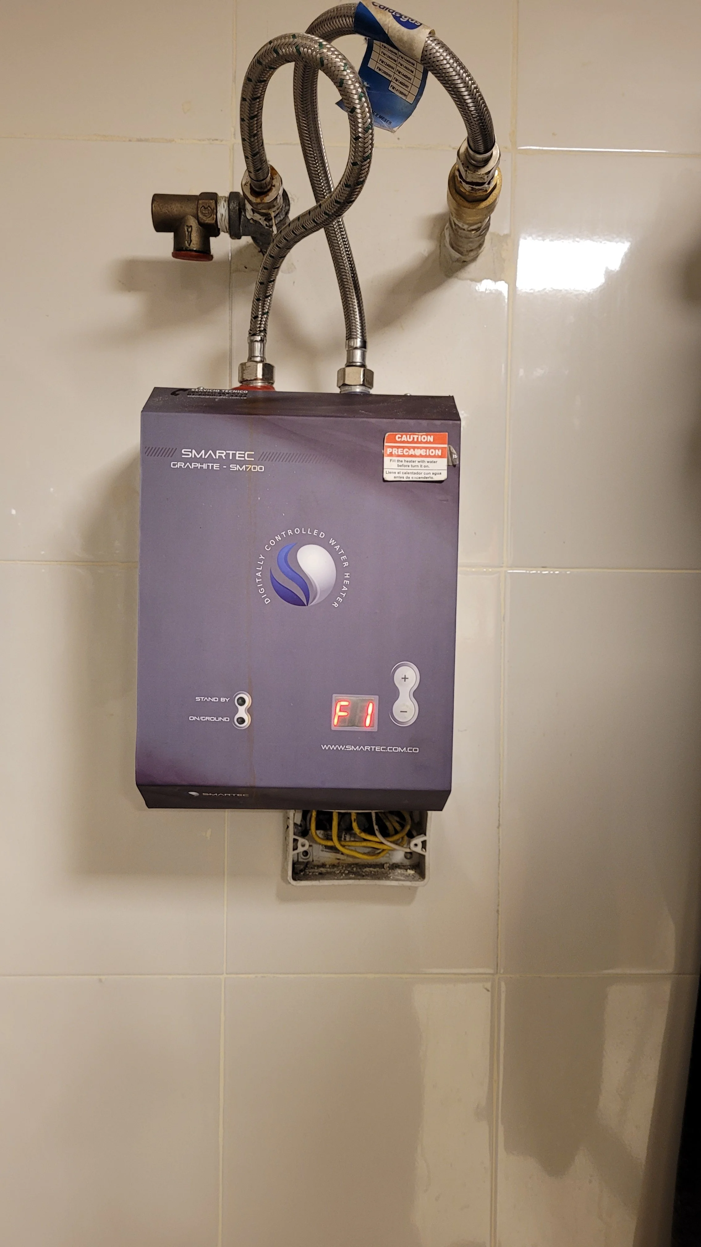 Calentador de agua controlado digitalmente, con pantalla que muestra F1, conectado a tuberías de agua en una pared de azulejos.