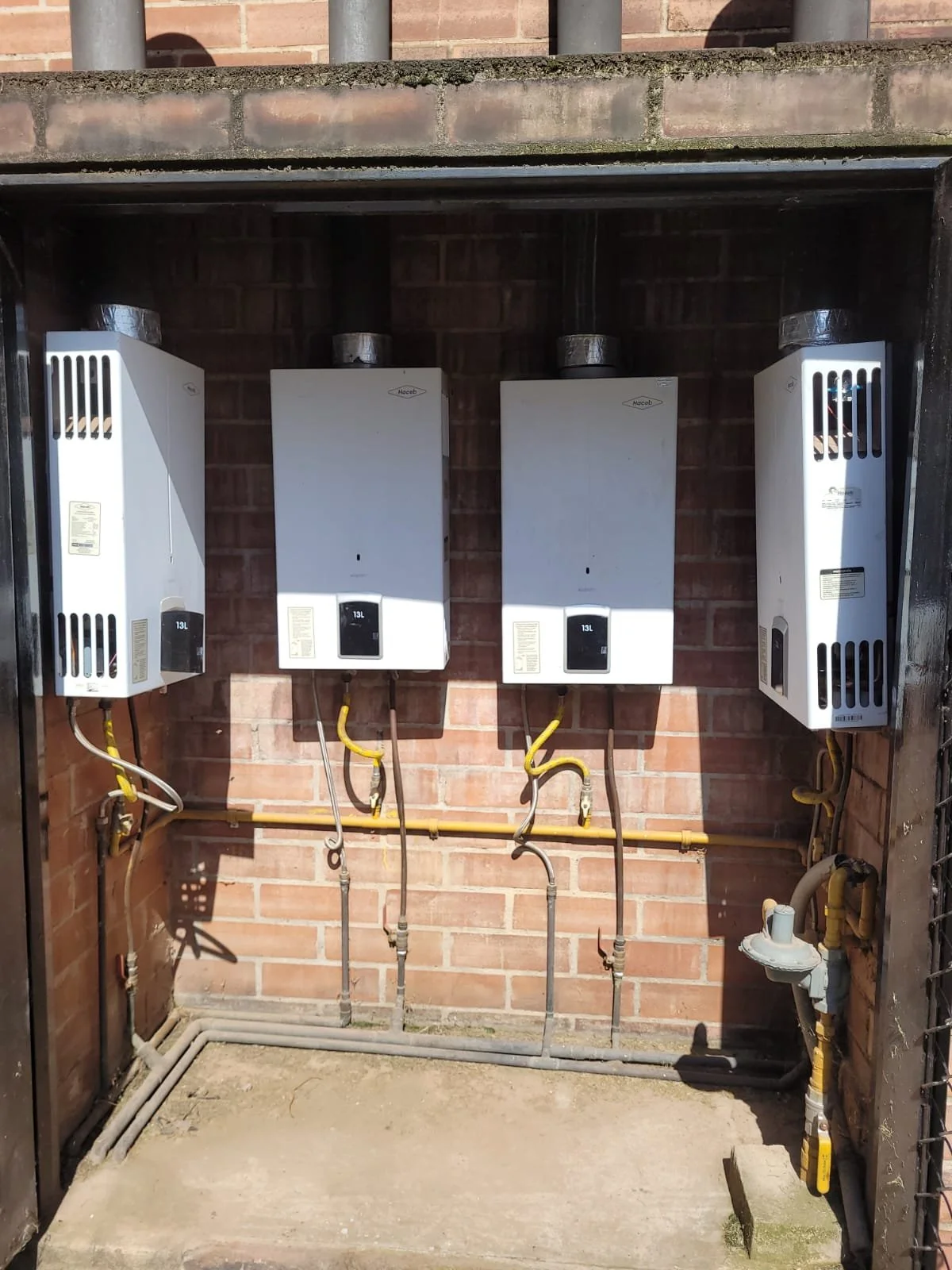 Tres calentadores de agua blancos instalados en una pared de ladrillos, con tuberías de metal y muestras de conexiones de gas, en un espacio al aire libre.