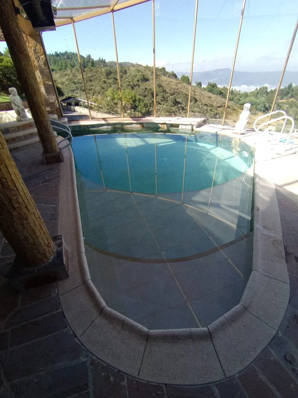 Piscina rodeada de ventanas con vistas a una colina y valle, decorada con estatuas blancas, en un espacio cerrado con paredes de piedra y estructura de madera.