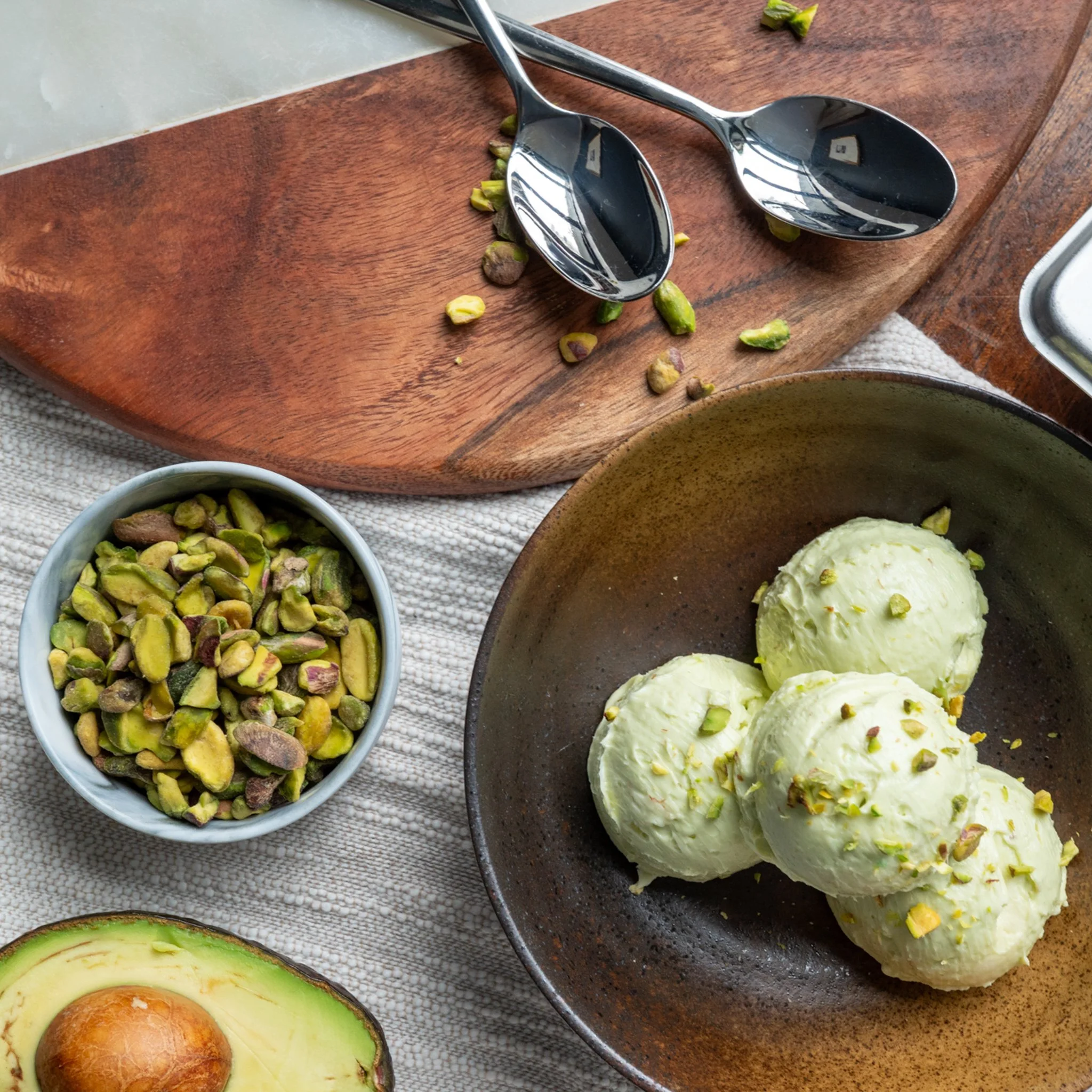 A bowl of three scoops of pistachio ice cream garnished with chopped pistachios, a small bowl of more chopped pistachios, a halved avocado with seed, and two metal spoons resting on a wooden board.
