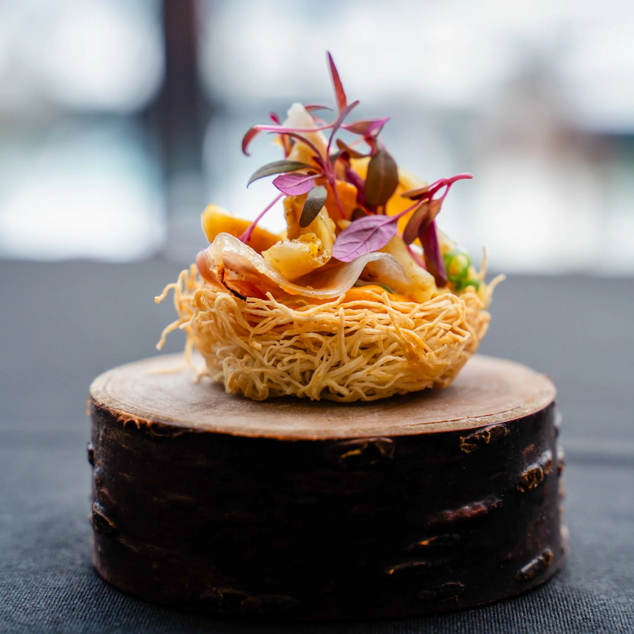A gourmet dish featuring a nest of crispy, golden fried noodles topped with assorted vegetables and garnished with purple microgreens, presented on a wooden stand.