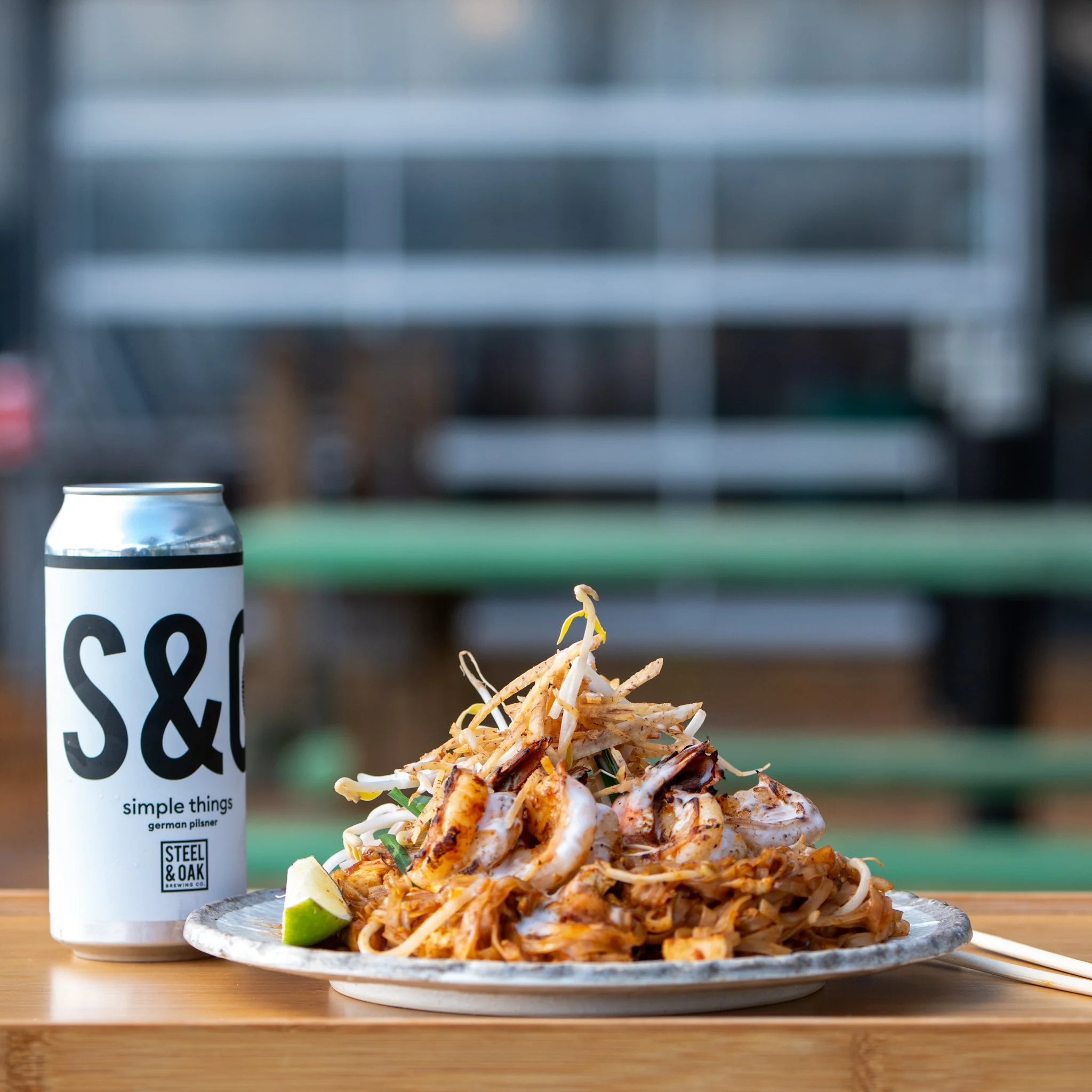 Plate of stir-fried noodles with vegetables and lime wedge, and a can of S&O beer with a modern urban background.