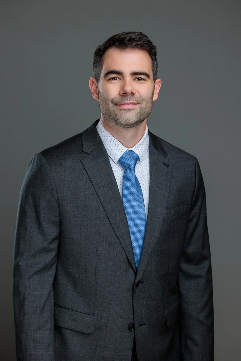 A professional man with dark hair and light beard wearing a dark gray suit, light blue tie, and white dress shirt with subtle pattern against a gray background.