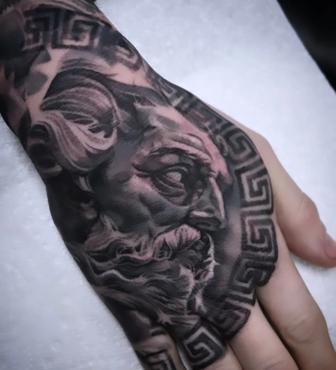 Tattoo of a Greek god or mythological figure with a beard, helmet, and a serious expression, surrounded by Greek key geometric patterns on a person's hand.