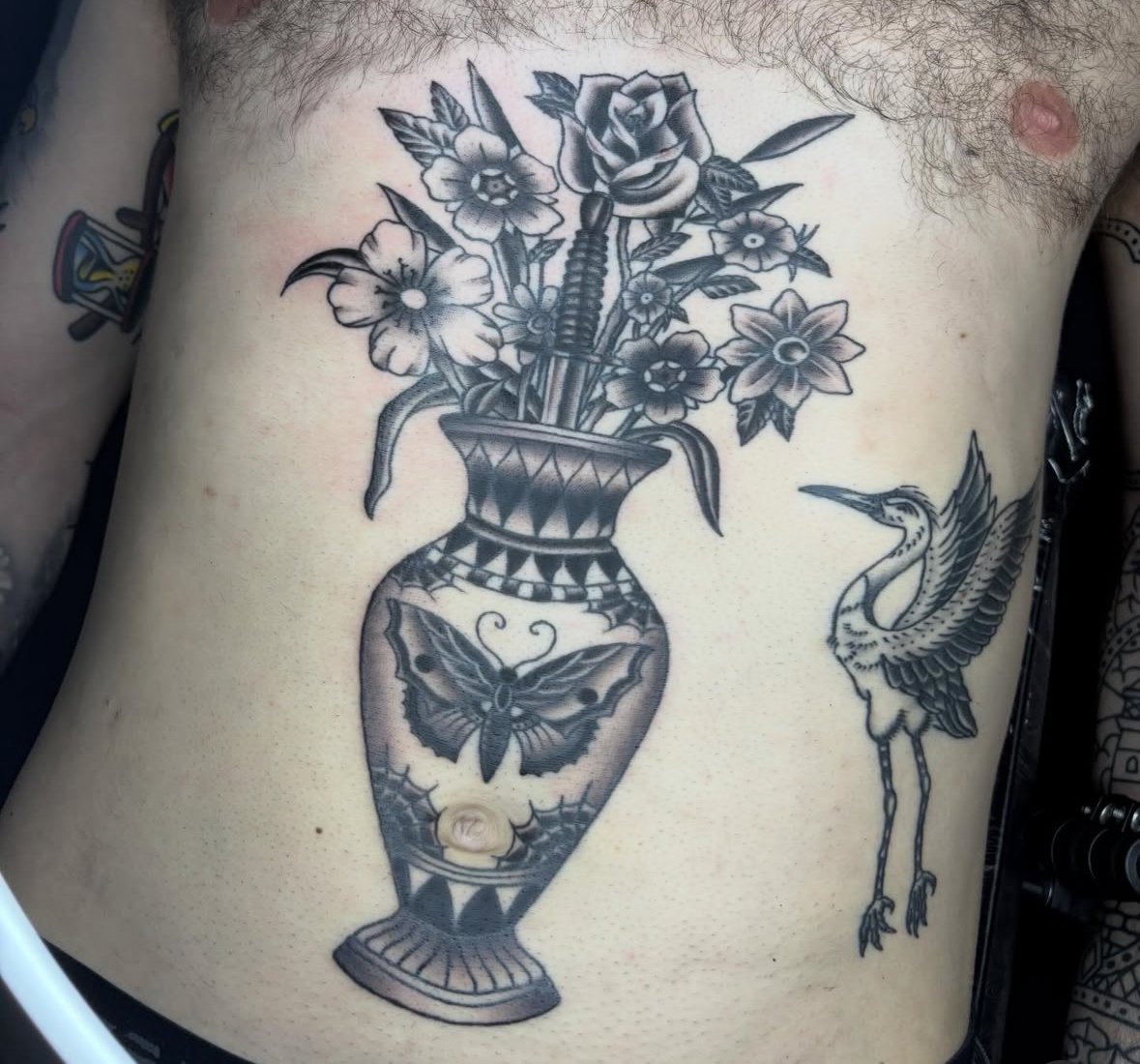A black and gray tattoo on a person's side featuring a vase with flowers and a dagger inside, alongside a heron bird.